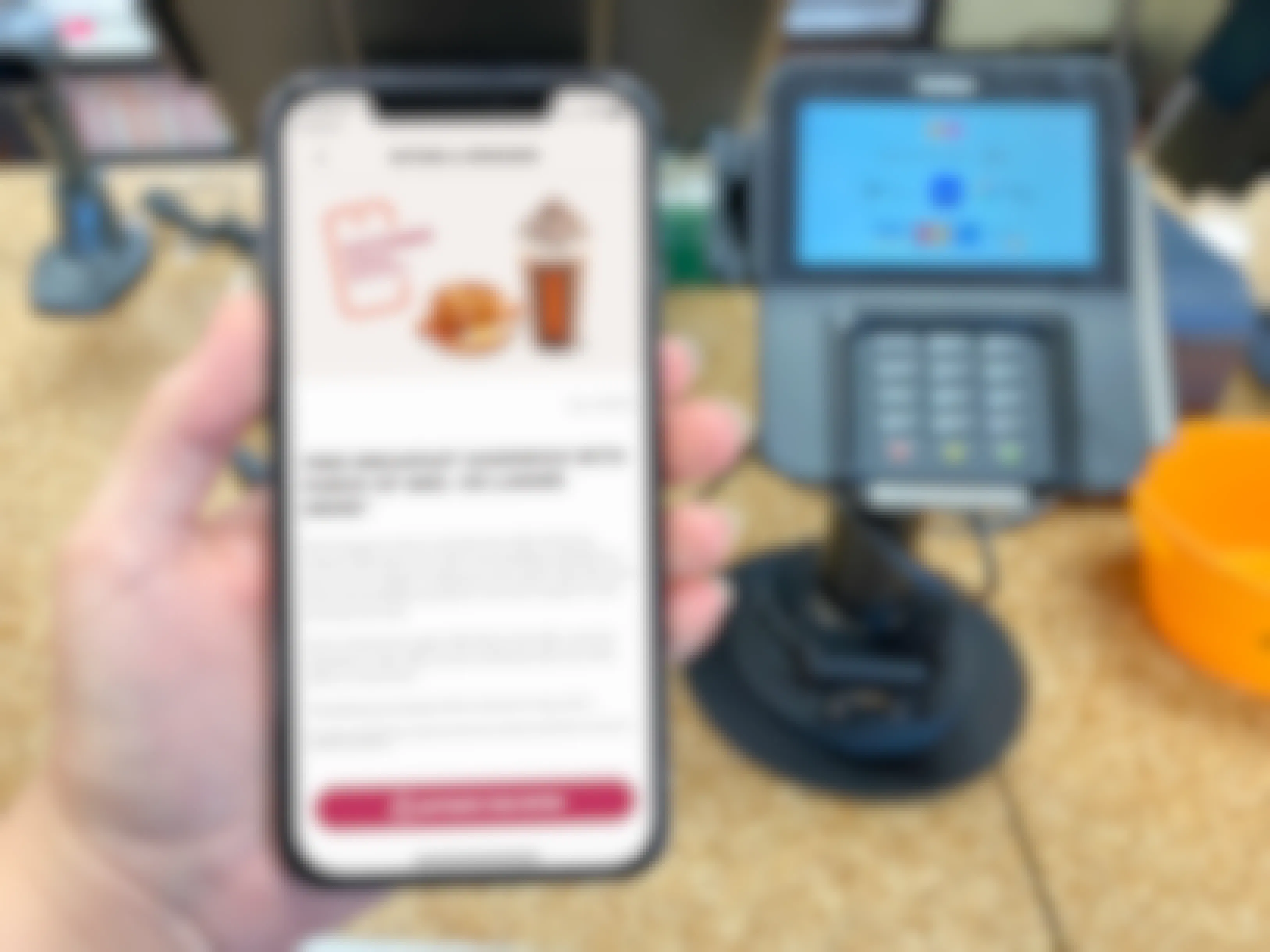 A person holding a cell phone at a Dunkin counter with an offer pulled up on the Dunkin Rewards app