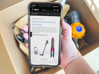 A person's hand holding a cellphone with dysons outlet website on screen with dyson vacuum in box in background
