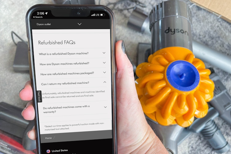 A person's hand holding a cellphone with dyson's outlet website on screen with dyson vacum on floor in background 