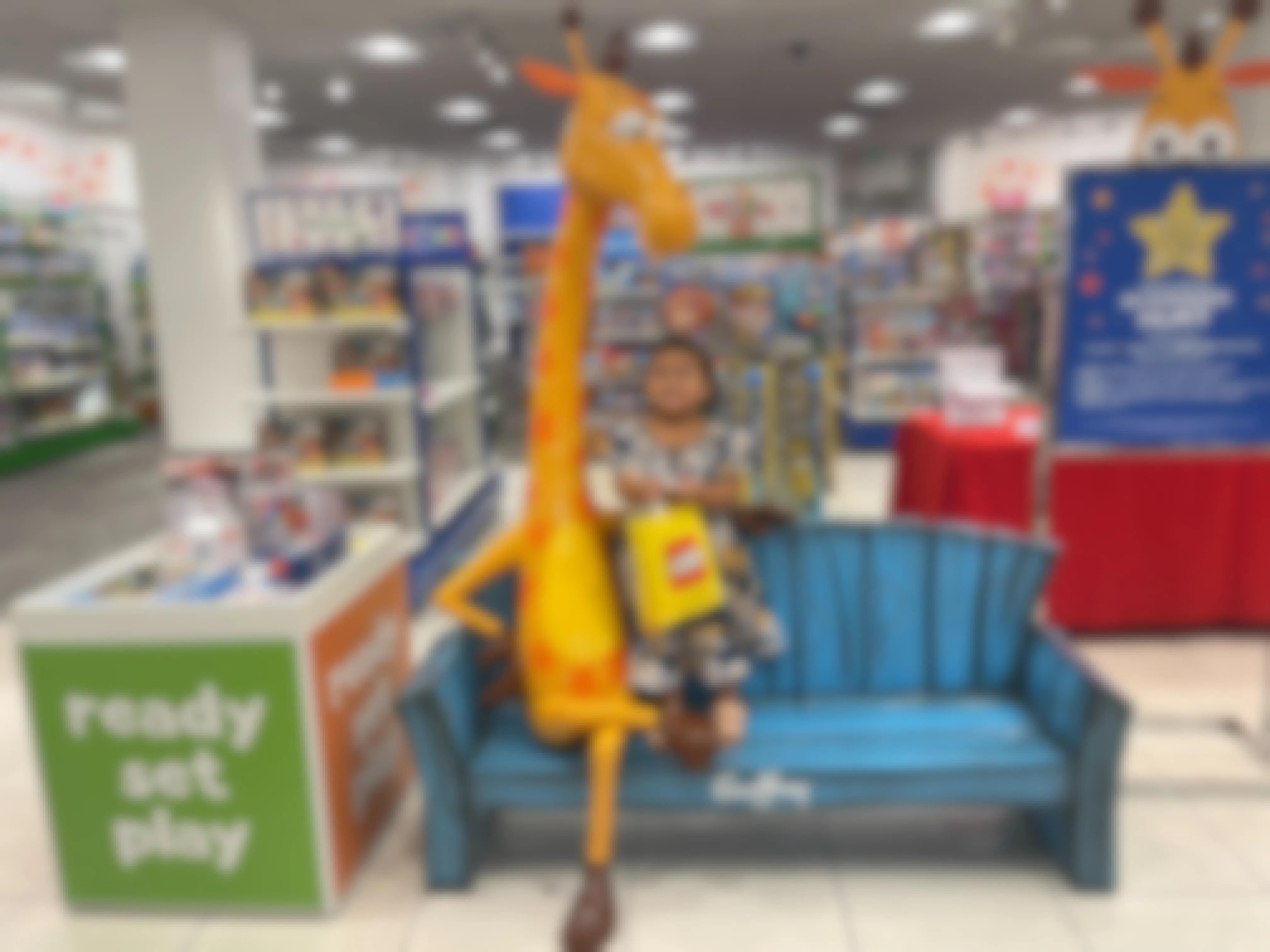 A small child sitting on a bench next to Toys R Us Geoffrey the giraffe in a store.