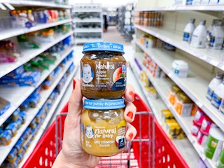 A variety of Gerber Natural babyhood held up by hand in front of shopping cart and store aisle.
