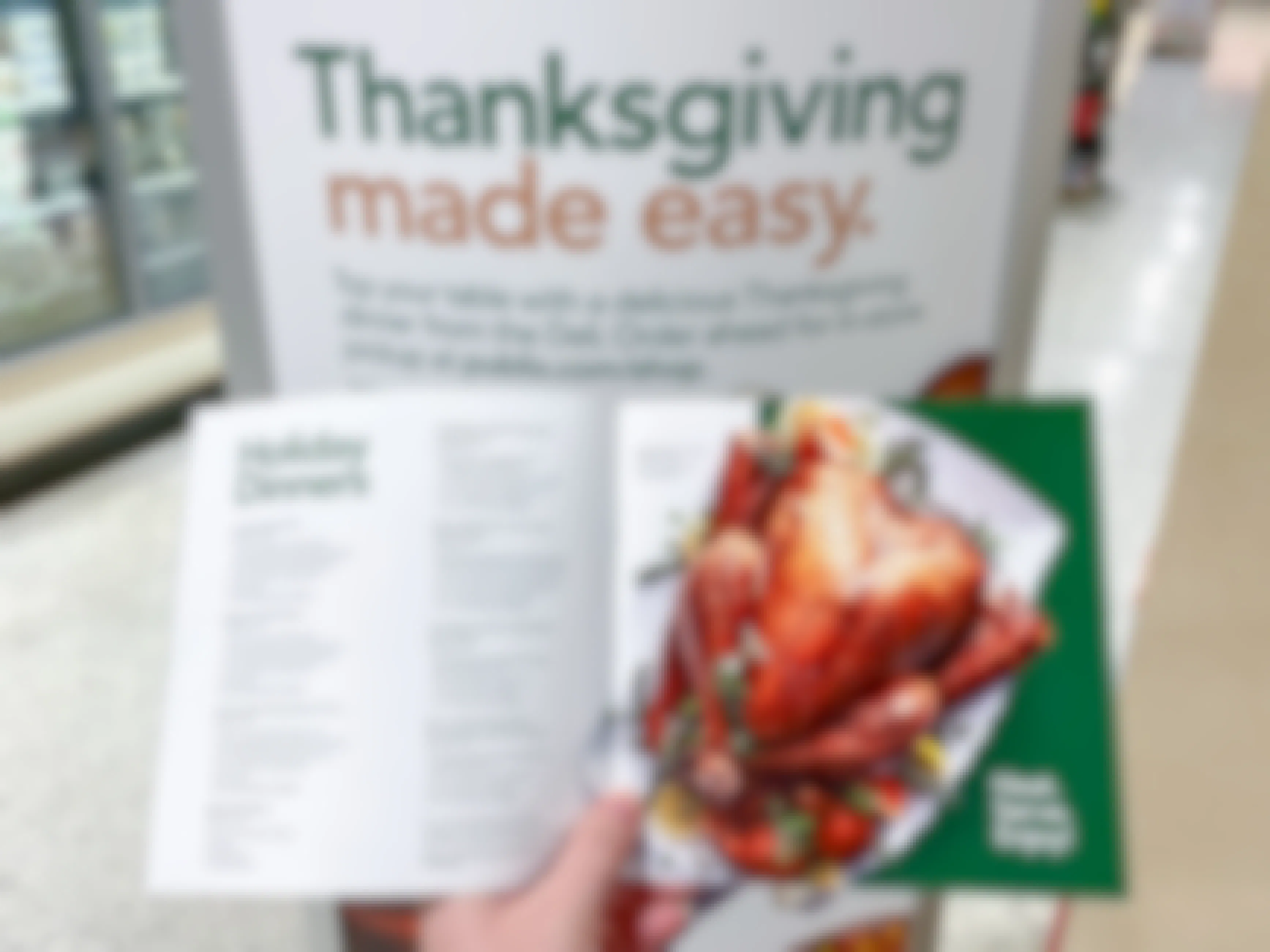 A person holding a booklet listing the Publix holiday dinner choices in front of a sign inside Publix that says, "Thanksgiving made easy. Top your table with a delicious Thanksgiving dinner from the Deli. Order ahead for in-store pickup at publix.com/shop