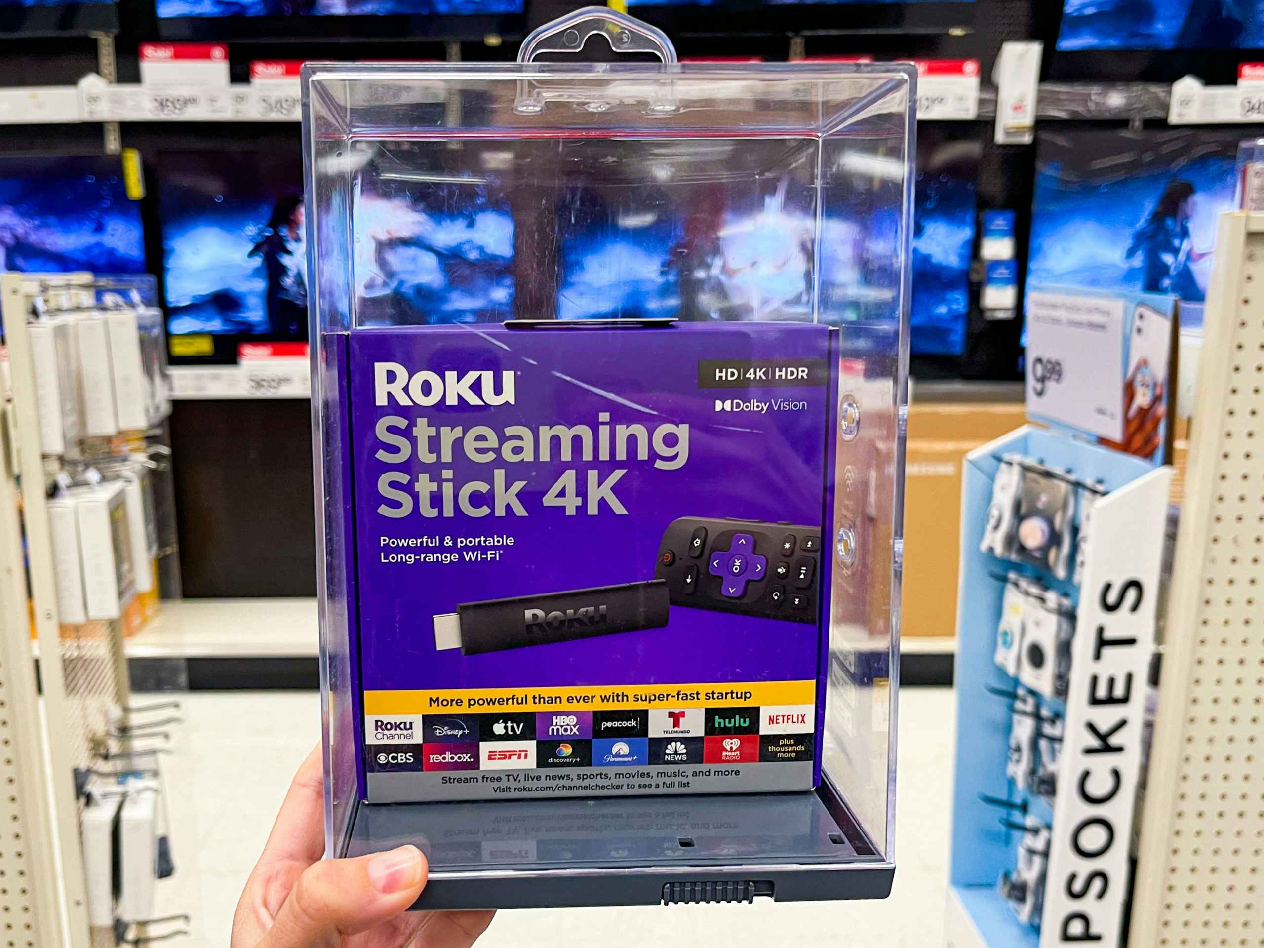 A Roku Streaming Stick 4k held out by hand in front of aisle.