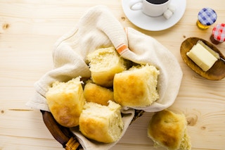 A basket with a cloth napkin filled with rolls on a table near a cup of coffee.