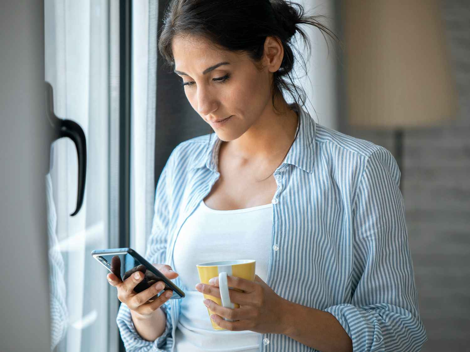 serious person holding coffee mug and looking at phone near window