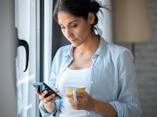 serious person holding coffee mug and looking at phone near window