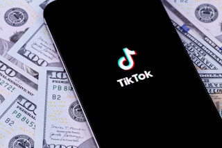 A phone displaying the TikTok app laying on a pile of cash.