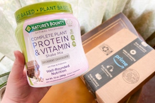 hand holding a tub of nature's bounty protein mix with an amazon prime box in the background