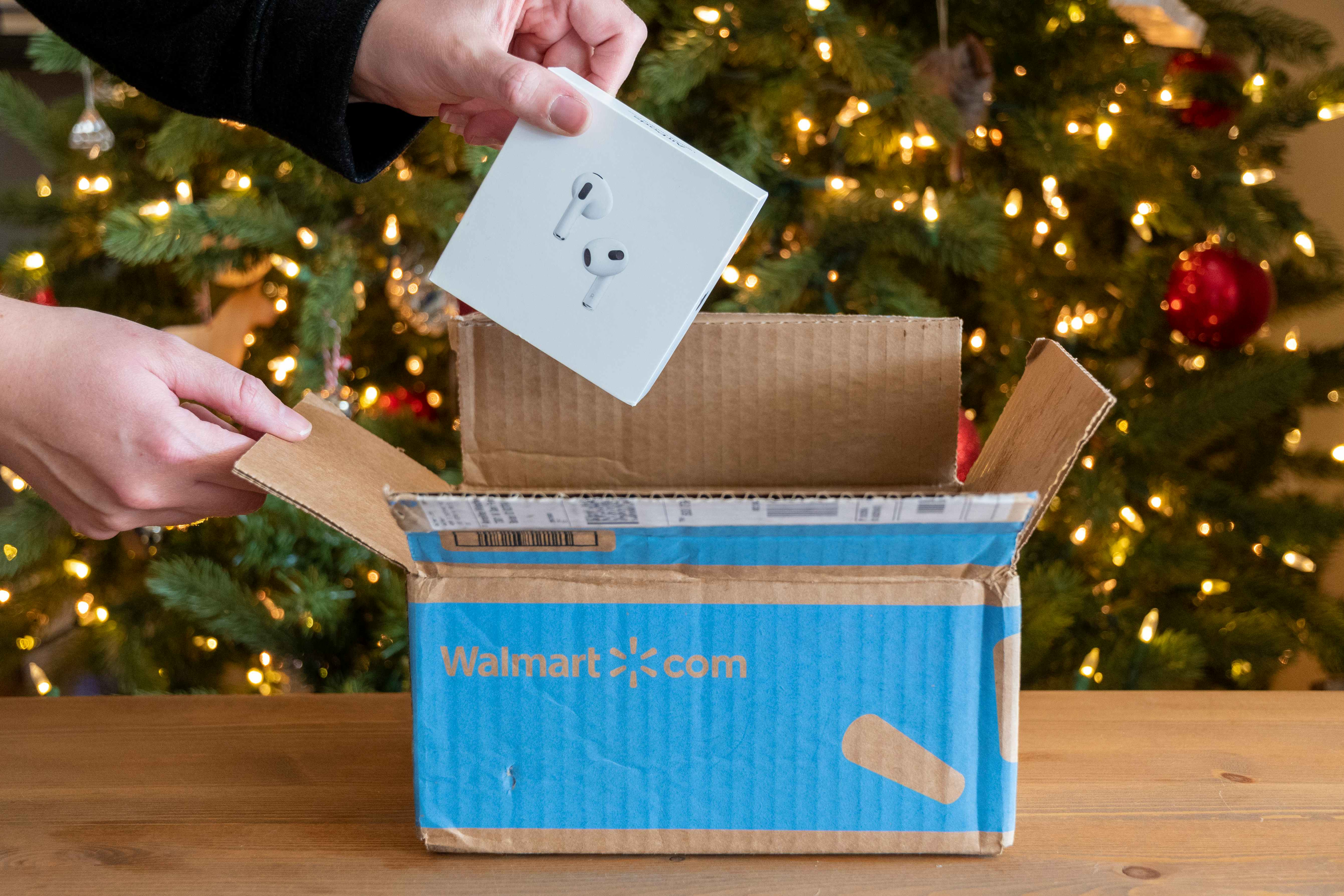 A woman's hand lifting Apple Airpods out of a Walmart shipping box in front of a lit Christmas tree