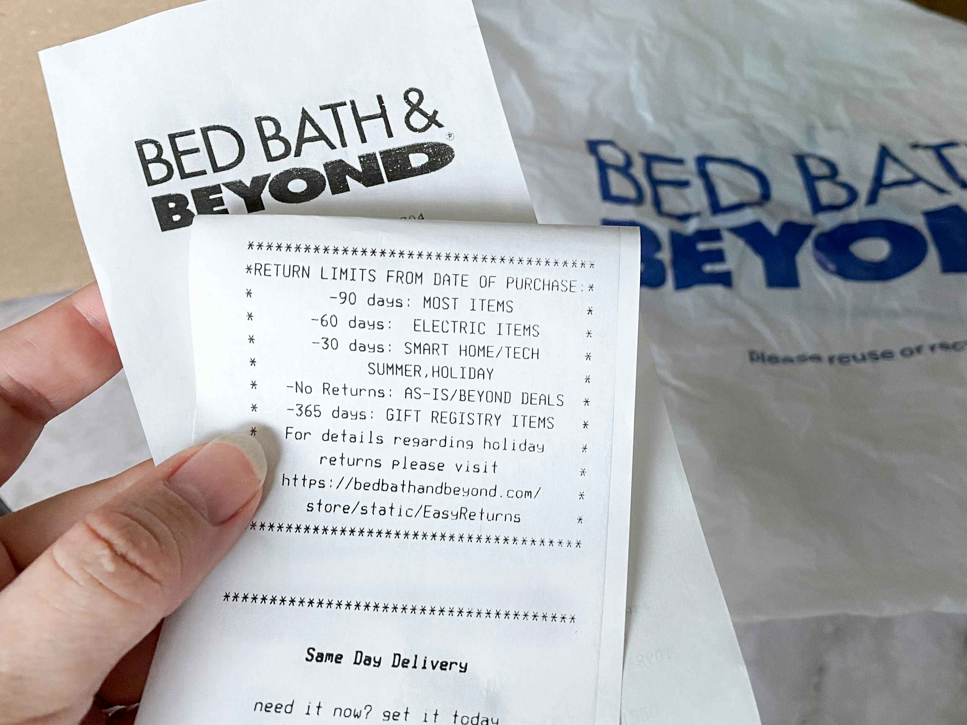A person holding a Bed Bath & Beyond receipt with the return policy showing and a shopping bag in the background