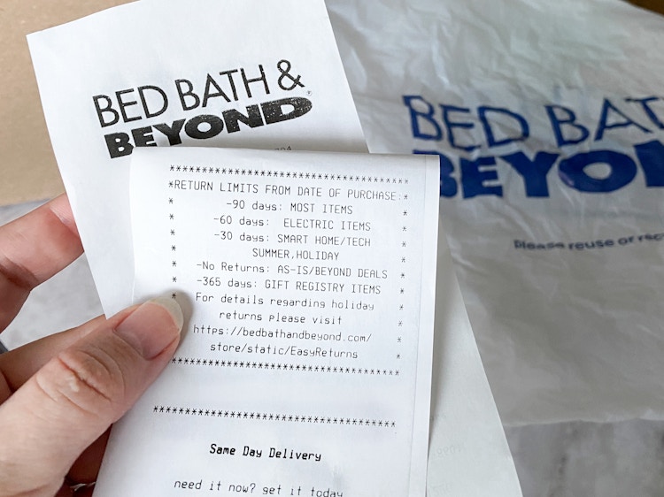 A person holding a Bed Bath & Beyond receipt with the return policy showing and a shopping bag in the background
