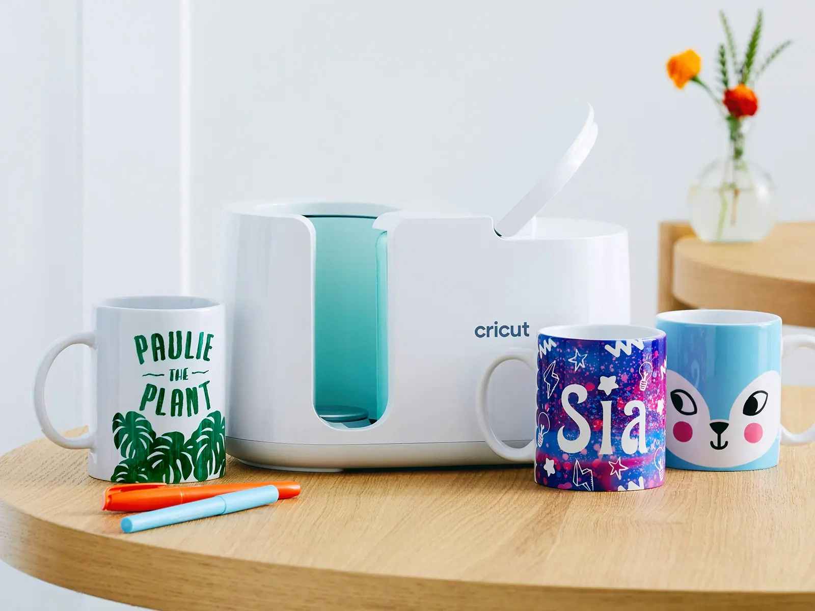A Cricut mug press on a table with some mugs that have been decorated using the mug press
