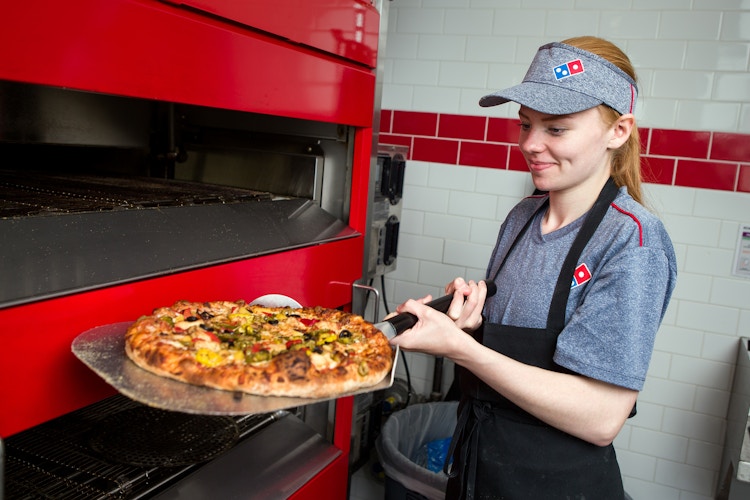 A Domino's employee taking a pizza out of the oven.