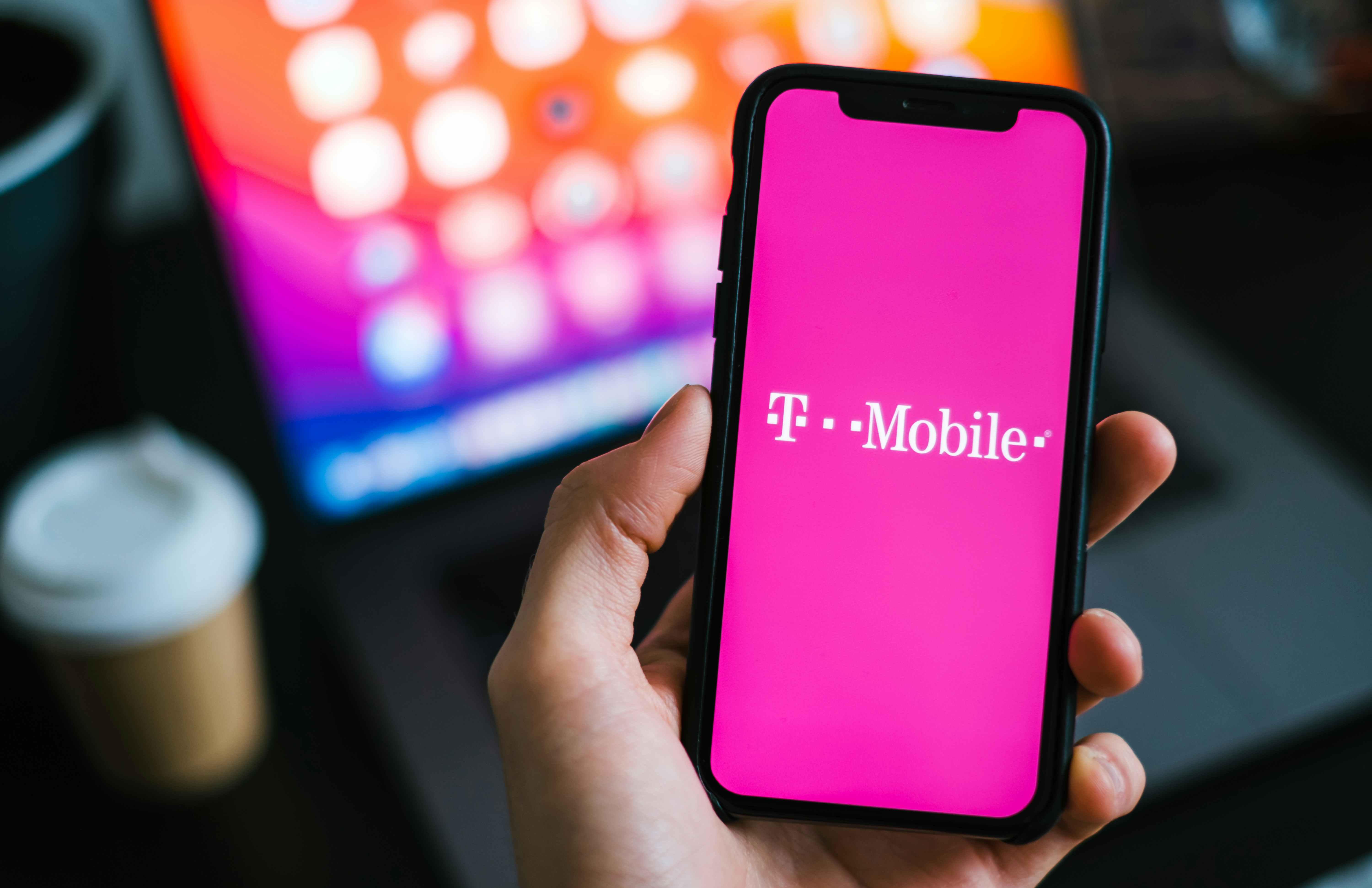 Person holding a phone with a t-mobile logo and laptop in the.background