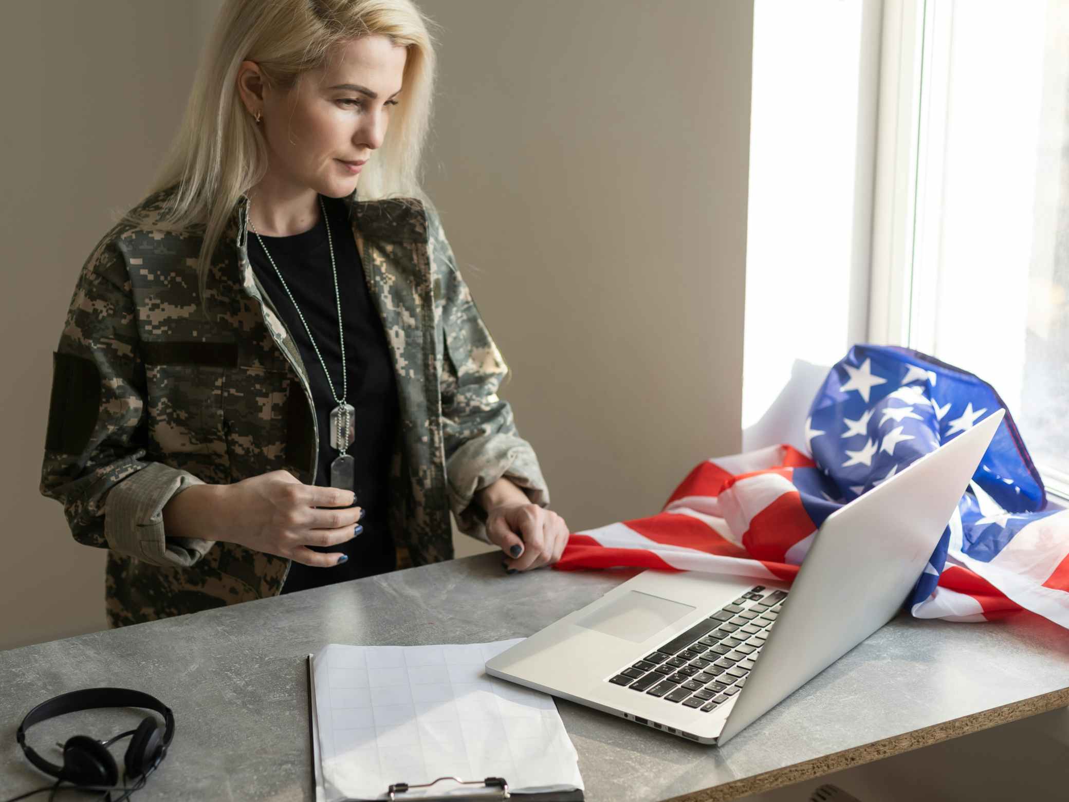 Female military looking at her laptop, on the counter there is a black headset and the american flag