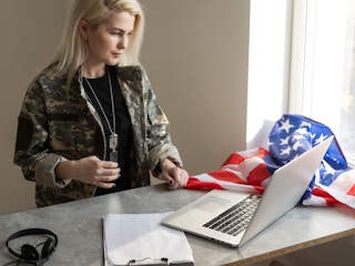 Female military looking at her laptop, on the counter there is a black headset and the american flag