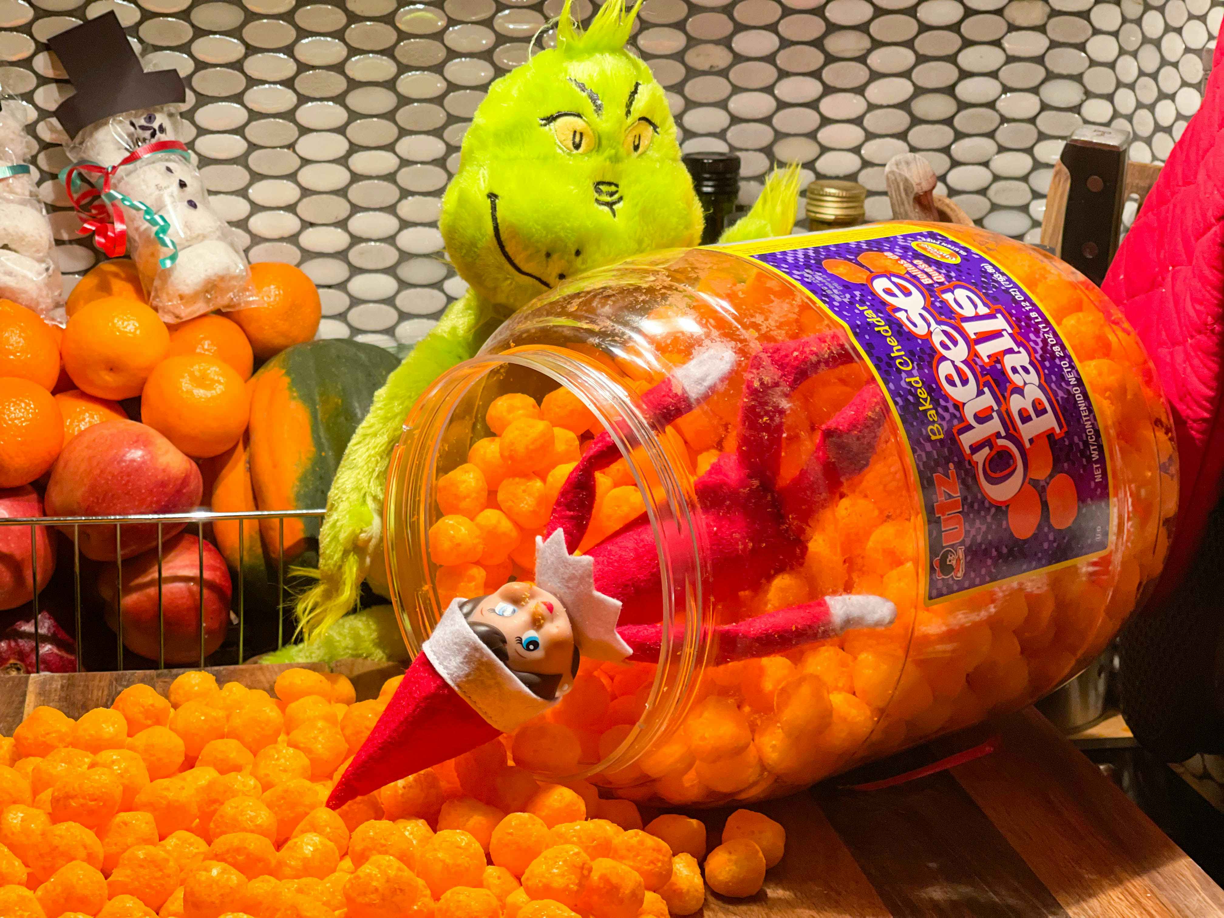 elf on the shelf doll inside a huge jar of cheese puffs with the puffs spilling out on counter and a grinch doll holding the container 