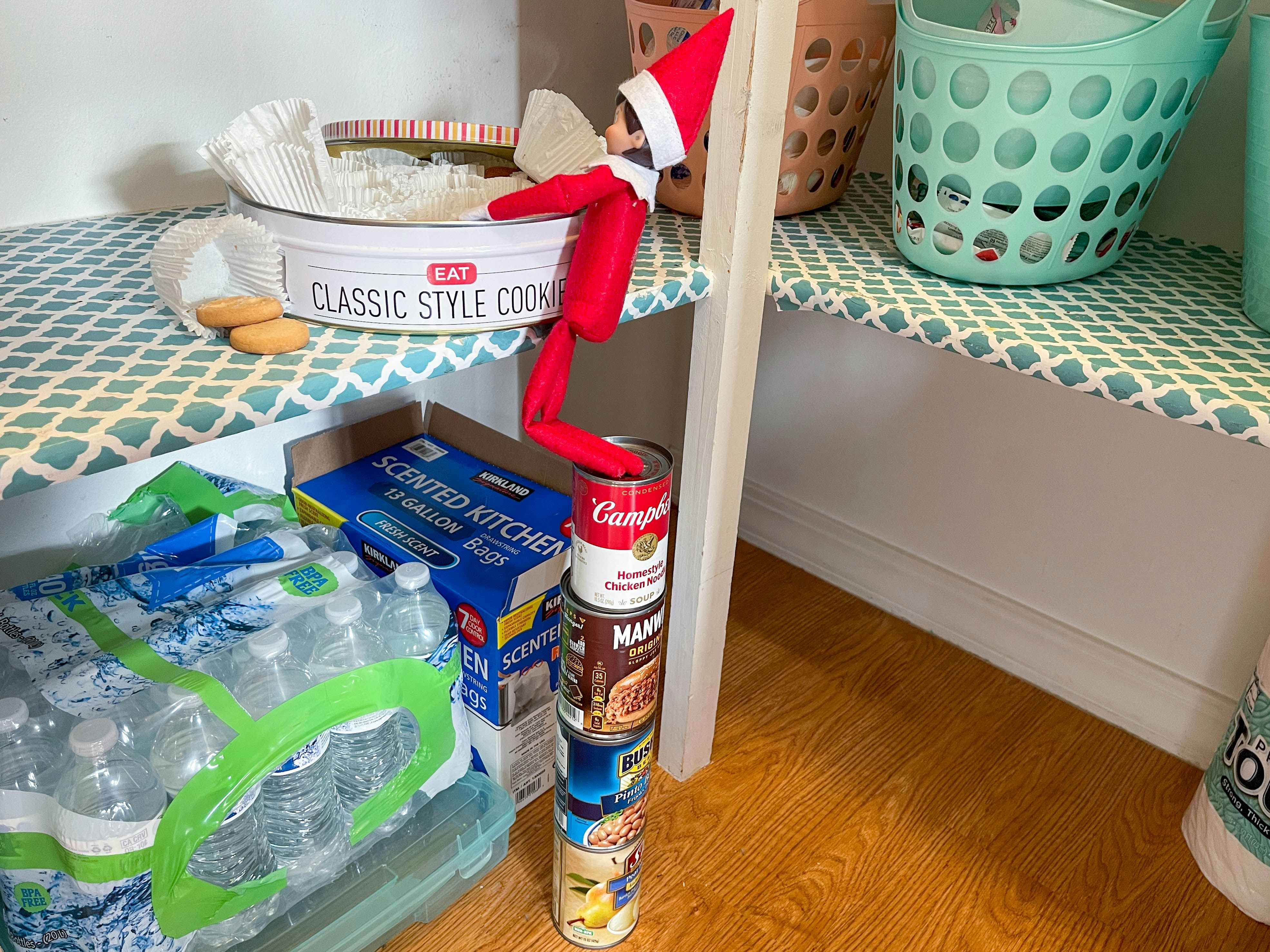 elf on the shelf doll standing on stacked cans getting into a tin of cookie in the pantry 