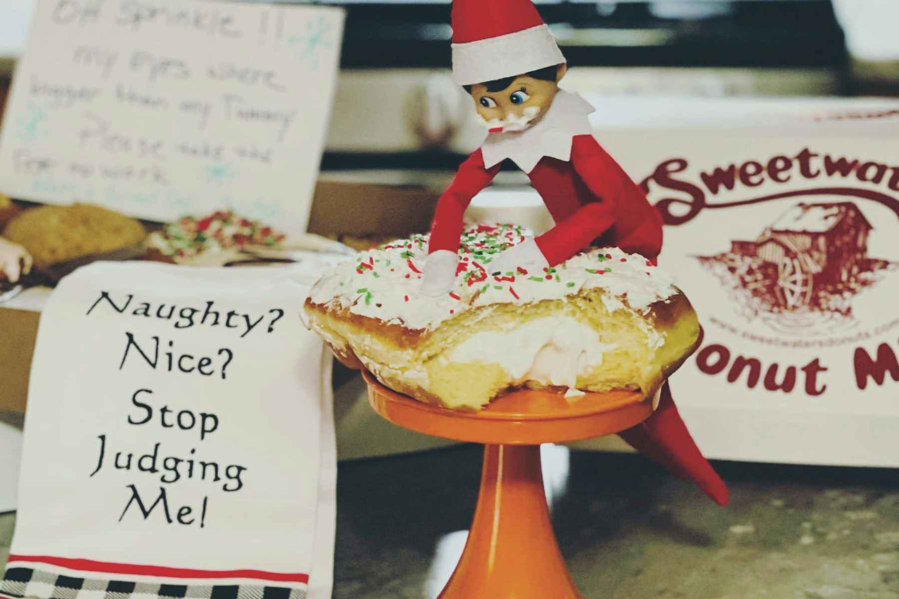 an elf on the shelf pretending to eat a donut next to a funny kitchen towel 