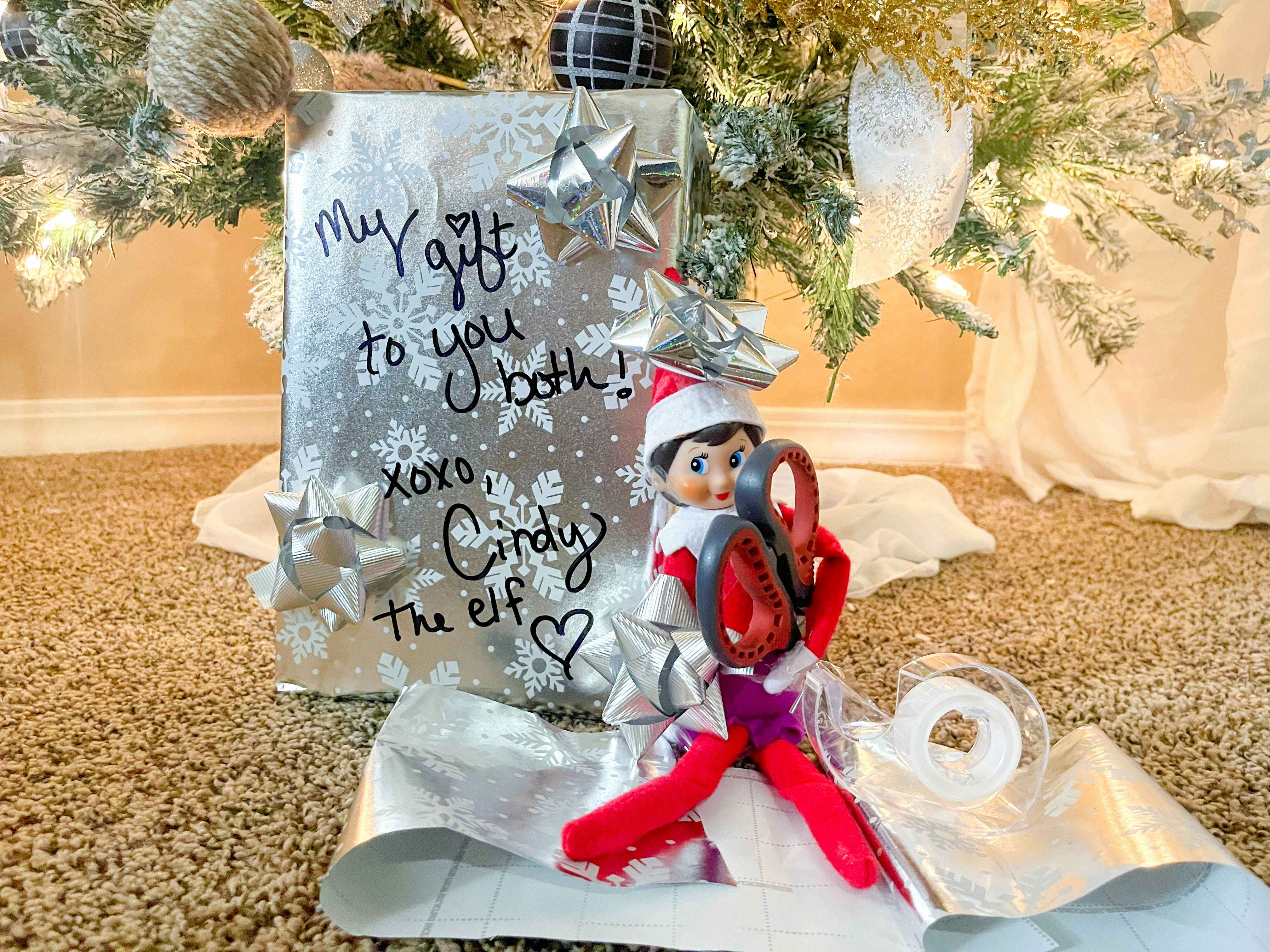 elf on the shelf doll sitting under a christmas tree next to a wrapped gift holding tape and scissors 