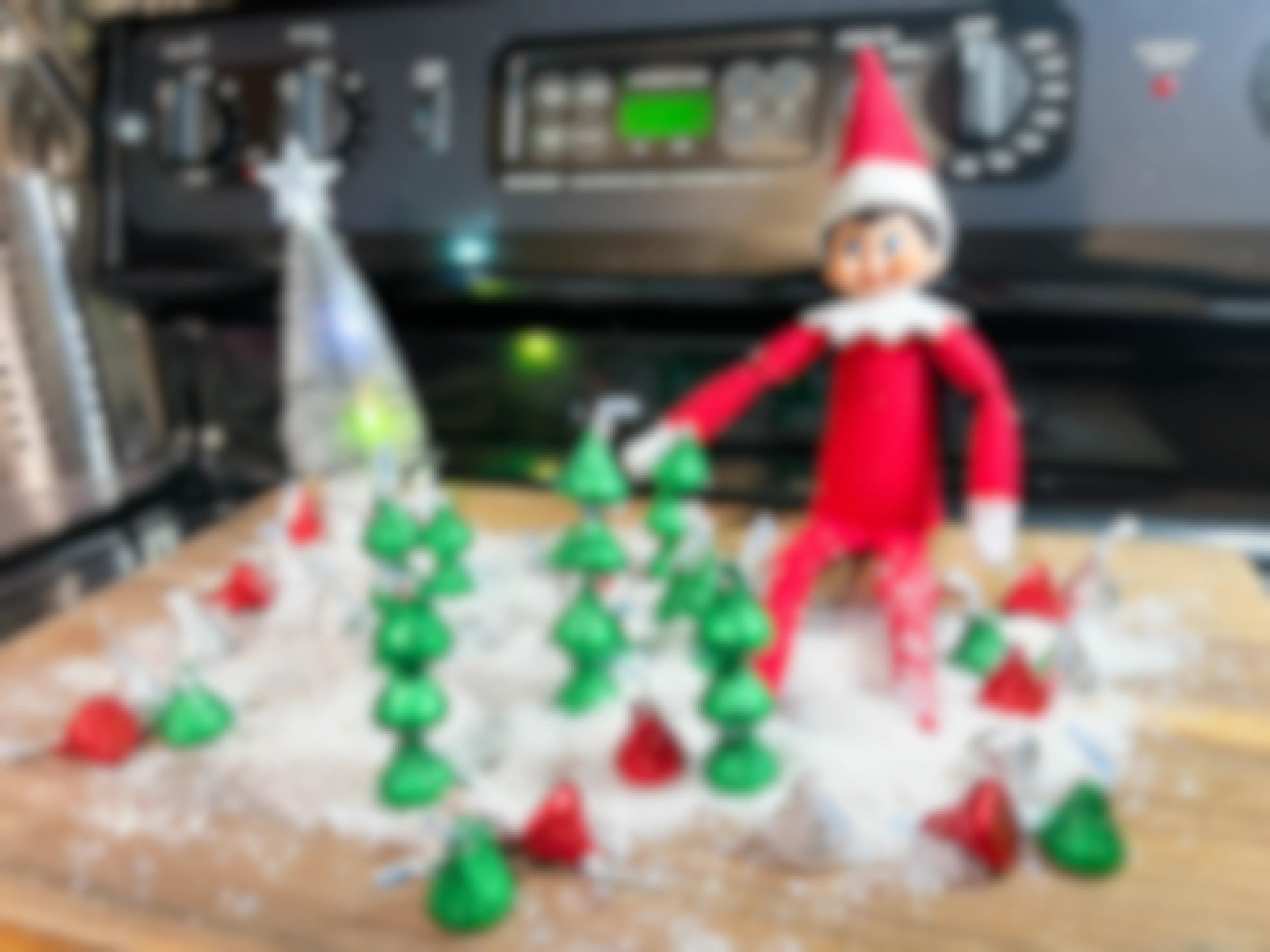 an elf on the shelf doll sitting on a cutting board with fake snow and christmas trees made out of hershey kisses