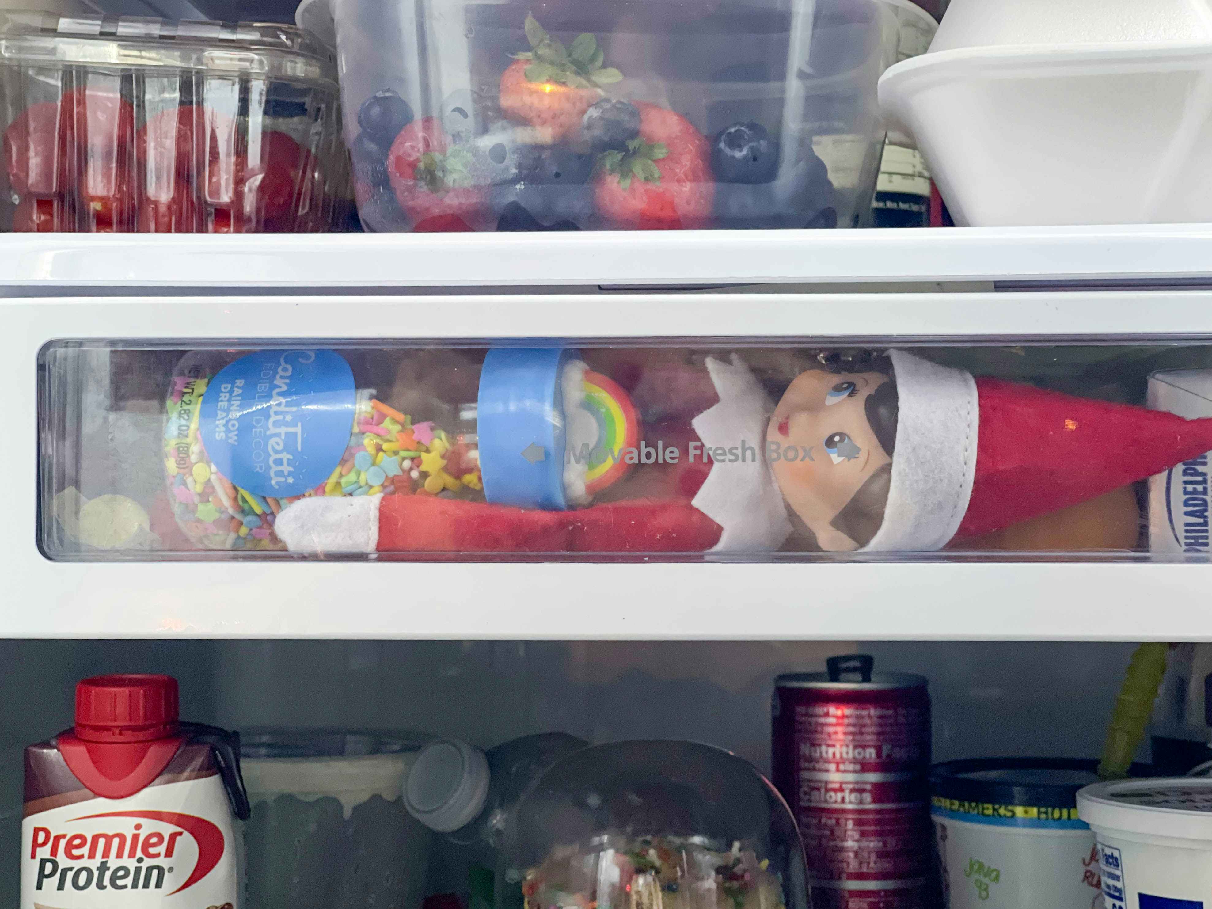 an elf on the shelf doll inside a fridge drawer holding sprinkles