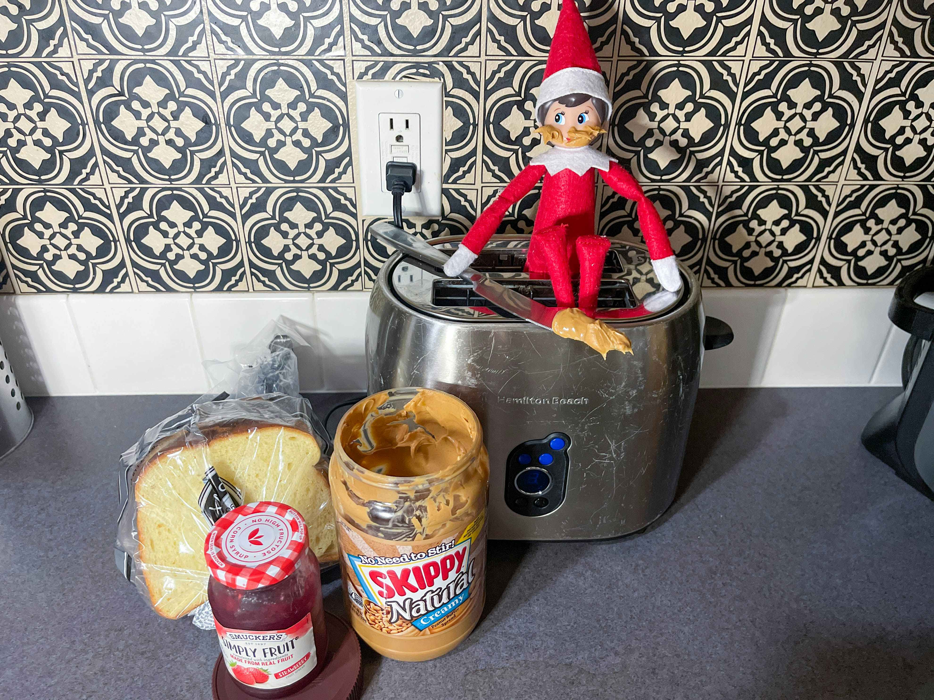 an elf on the shelf doll sitting on a toaster with a knife loaded with peanut butter and peanut butter on its face with bread, jam, and jar of peanut butter next to the toaster 