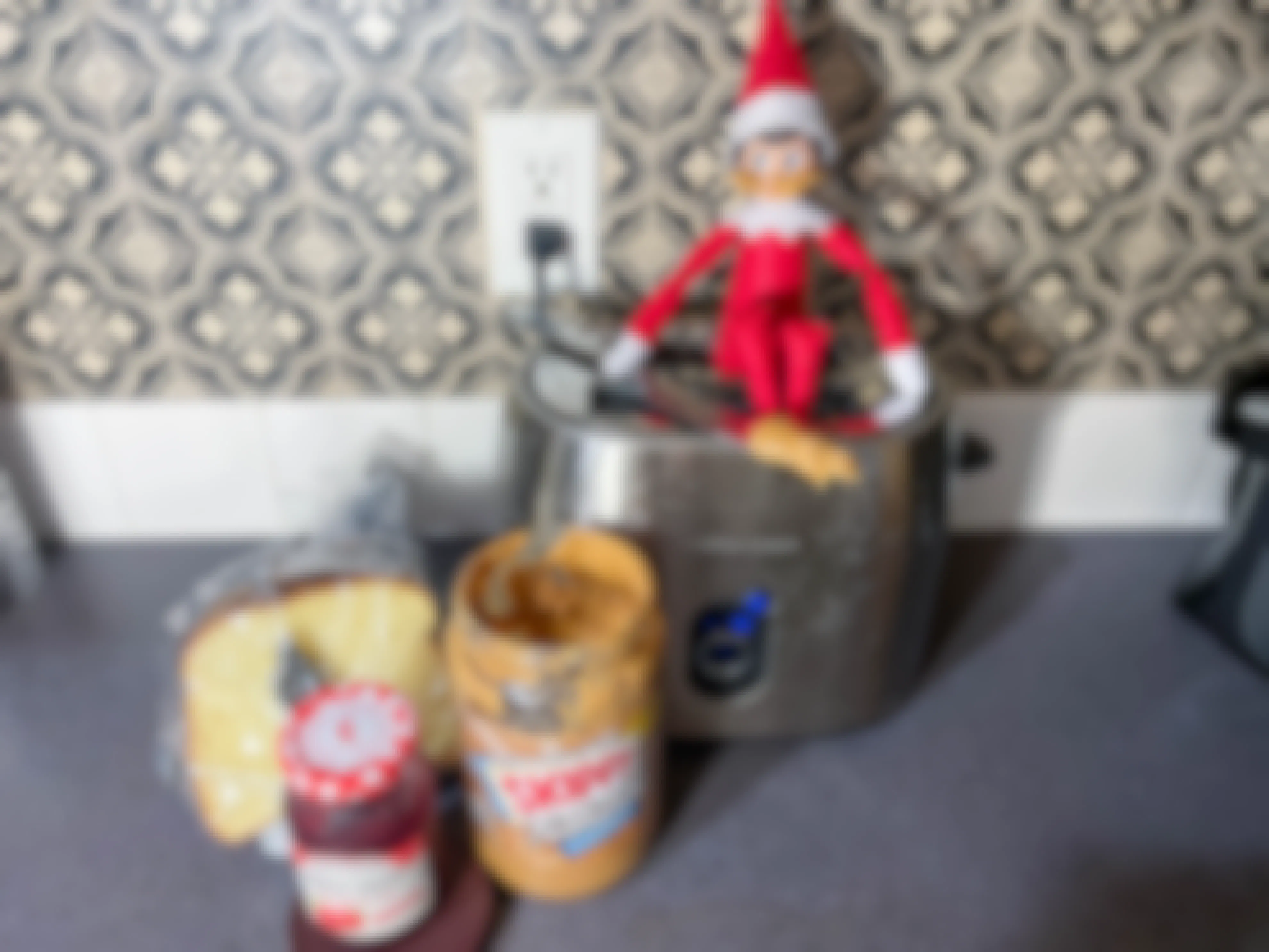 an elf on the shelf doll sitting on a toaster with a knife loaded with peanut butter and peanut butter on its face with bread, jam, and jar of peanut butter next to the toaster 