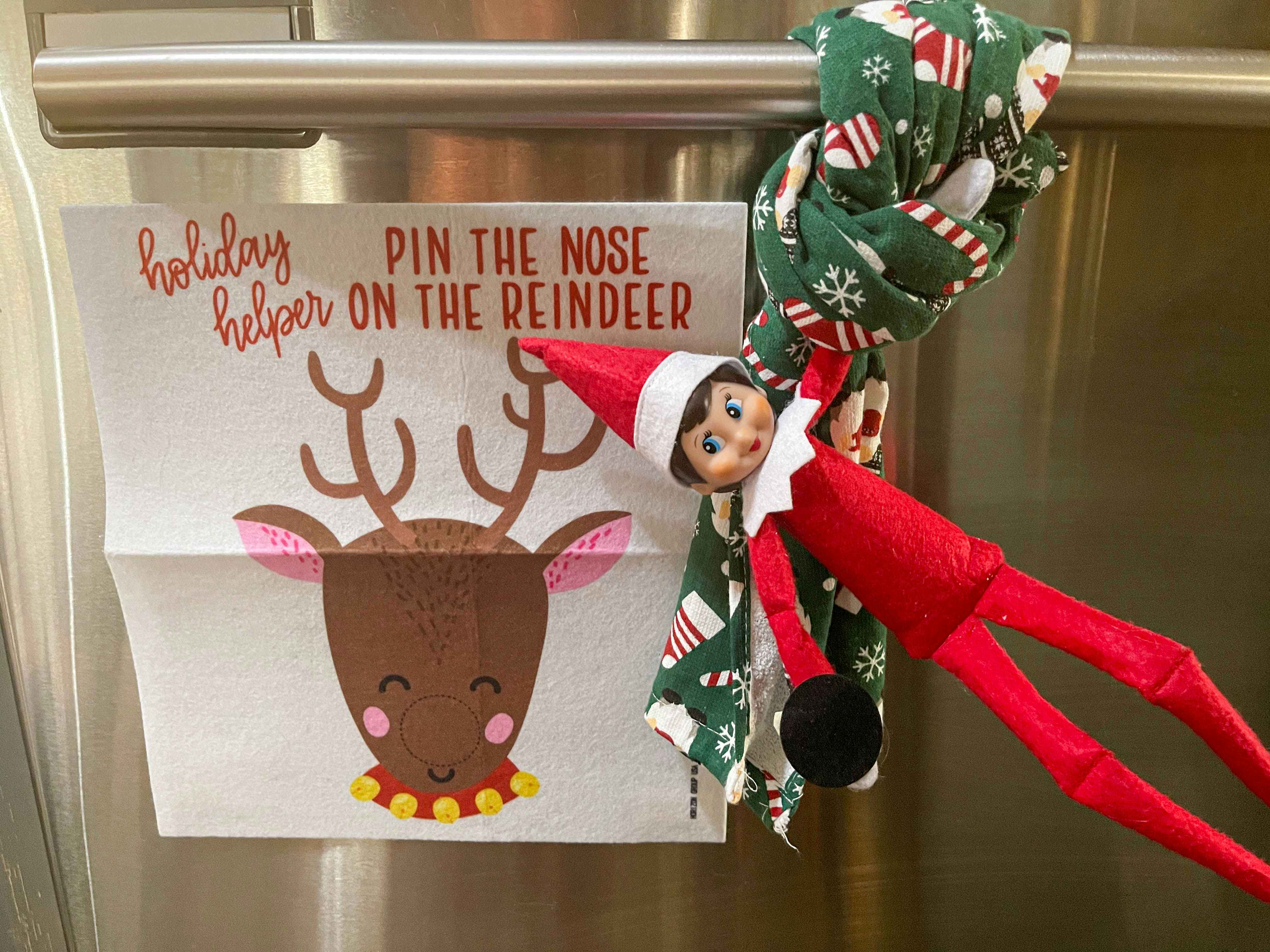 an elf on the shelf doll playing pin the nose on the reindeer holding on a kitchen towel