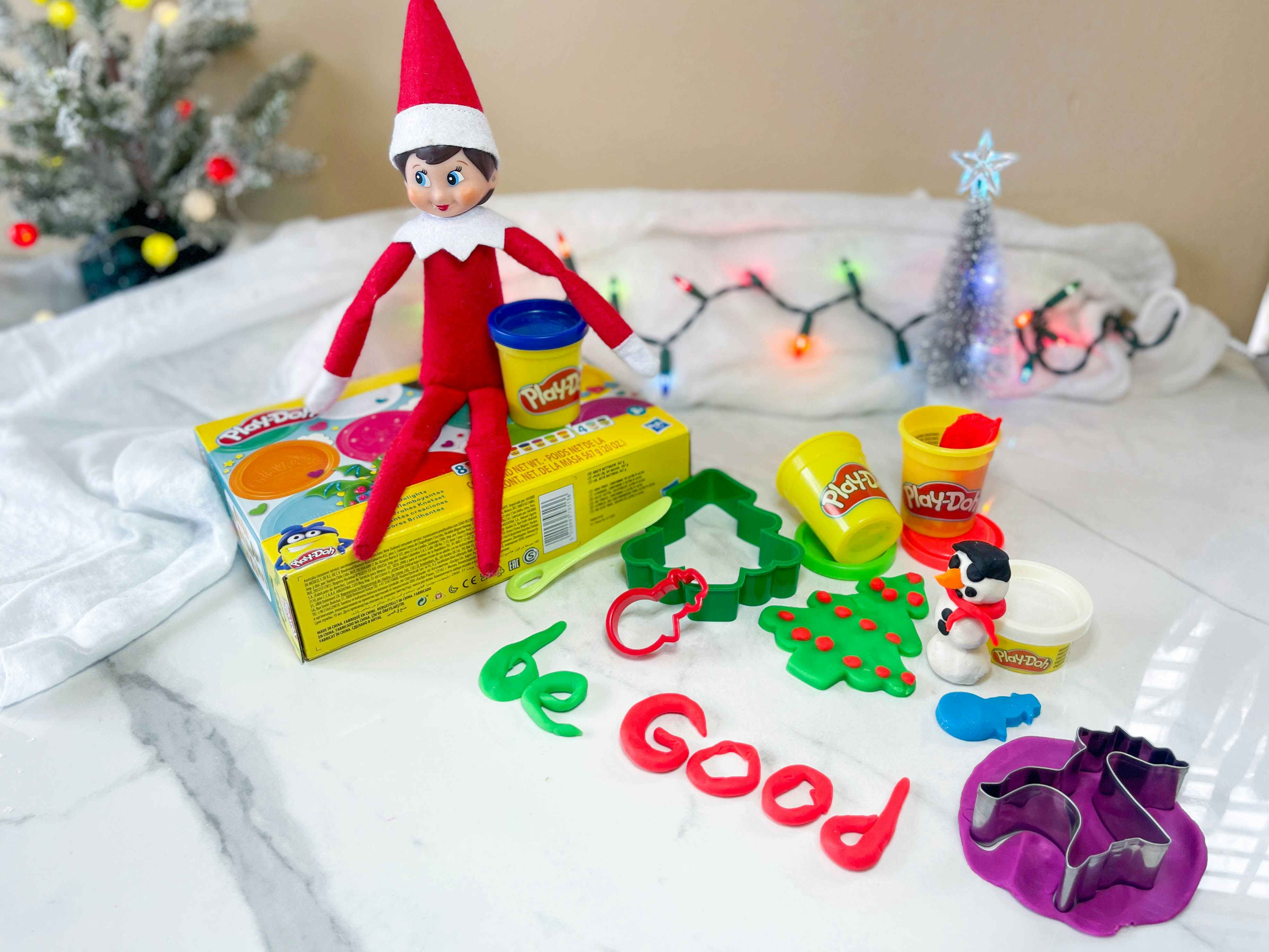 elf on the shelf doll sitting on top a playdoh box next to playdoh spelling out be good and other holiday playdoh supplies