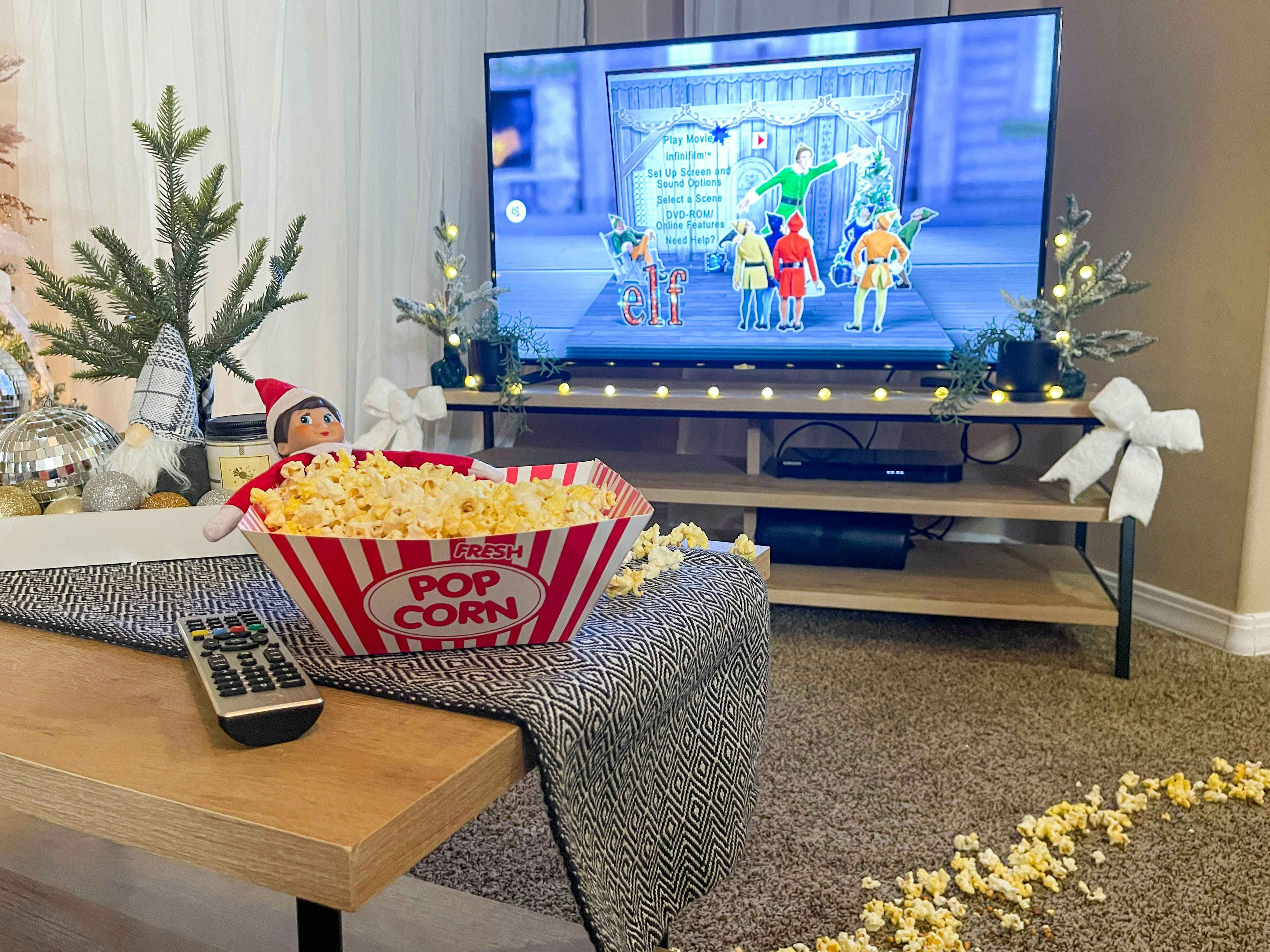elf on the shelf doll laying inside a tray of popcorn in front of a tv with elf movie on screen 