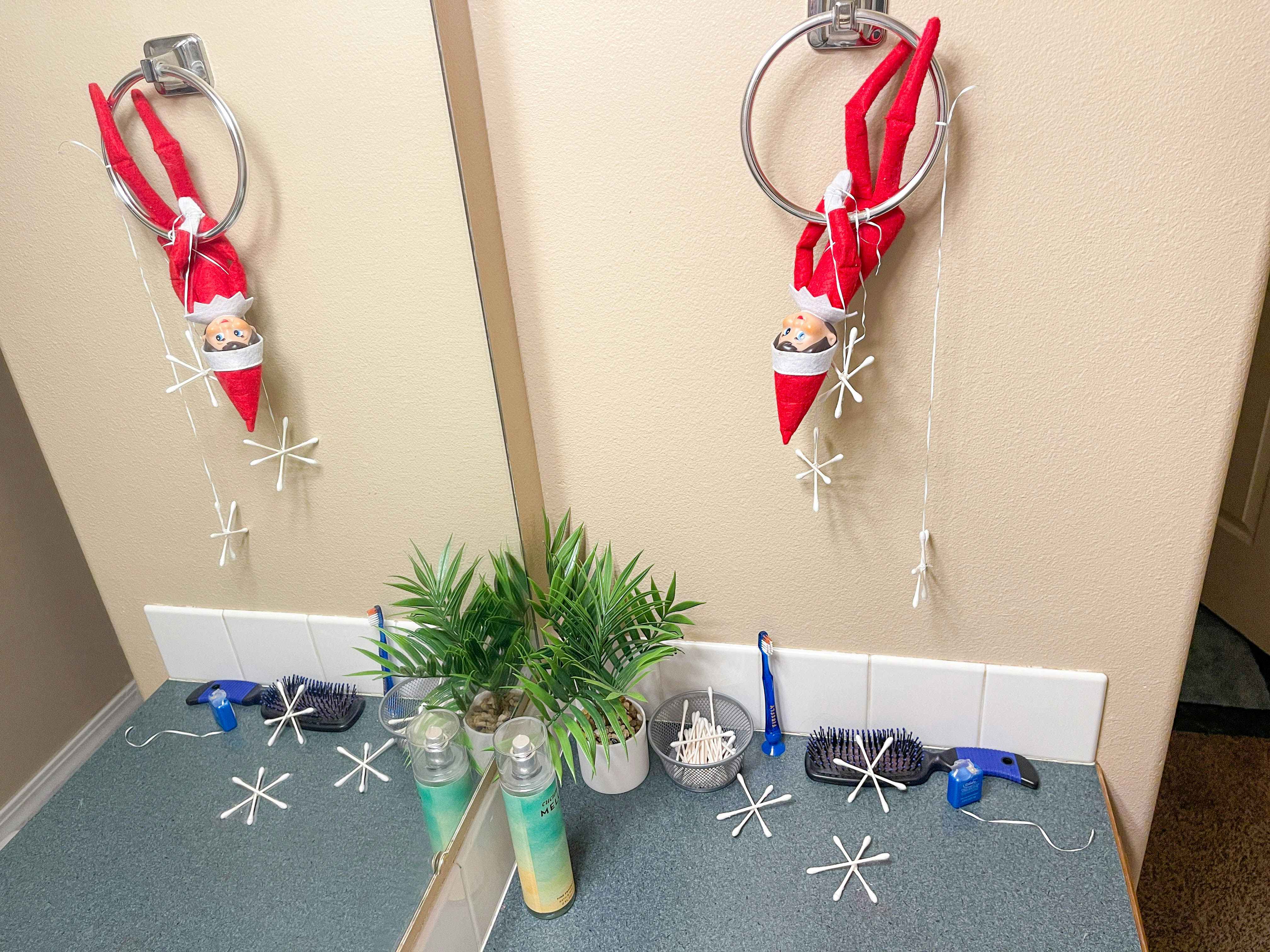 an elf on the shelf doll hanging upside down on towel ring in bathroom with qtip snowflakes around counter