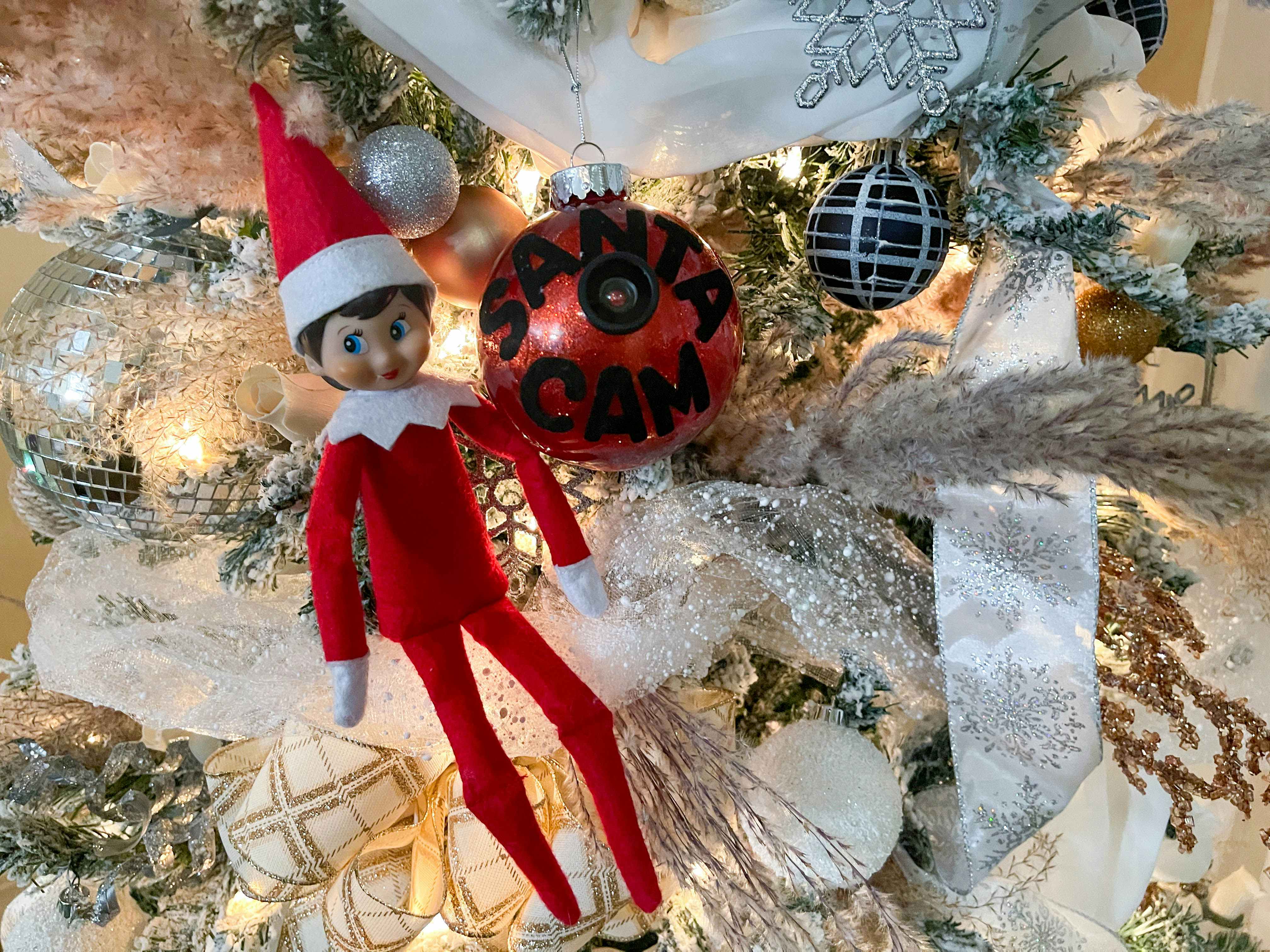 an elf on the shelf sitting next to a diy santa cam ornament hanging in a tree 