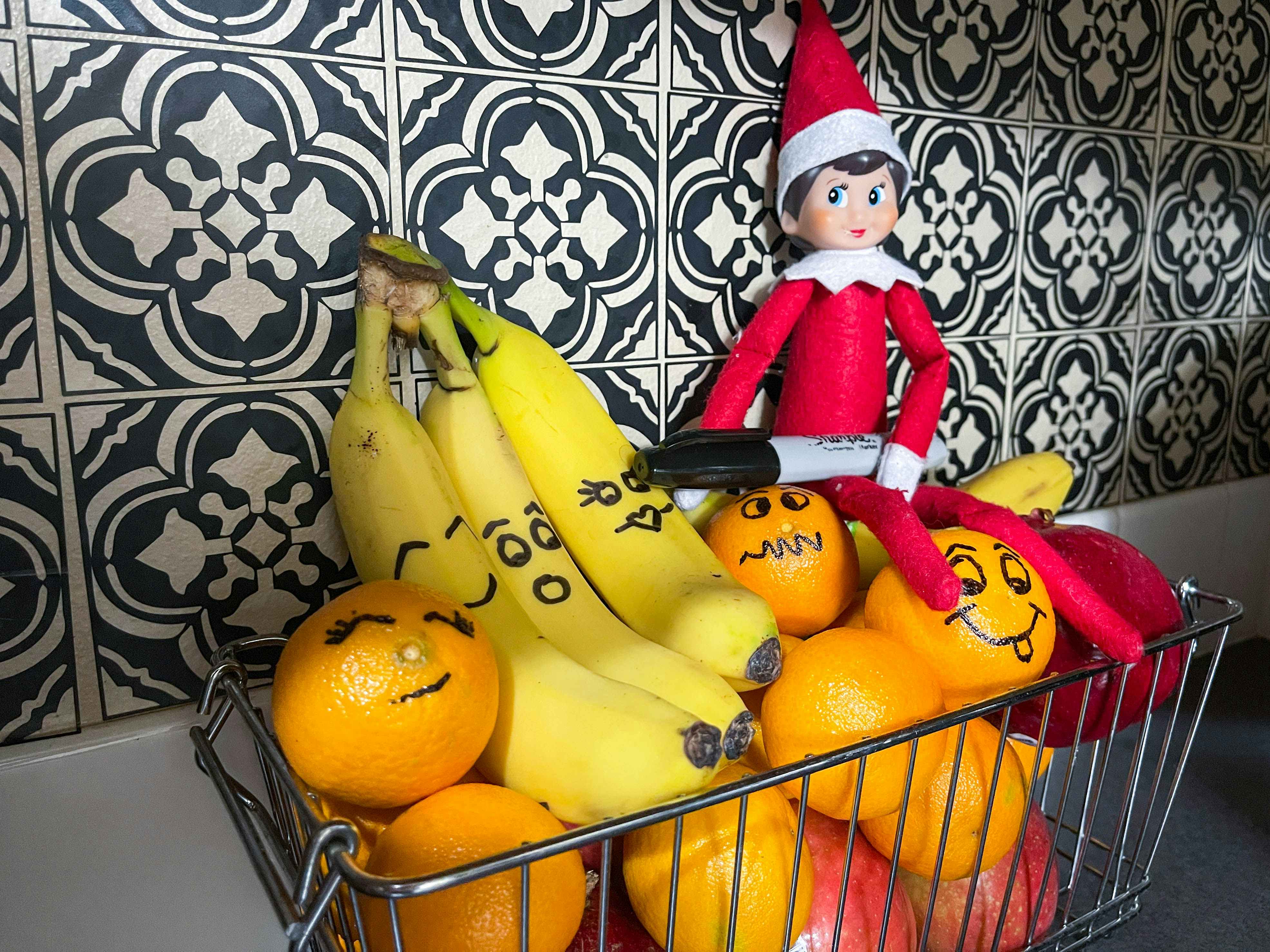 an elf on the shelf doll holding sharpie with faces marked on bananas and oranges in a basket of fruit 