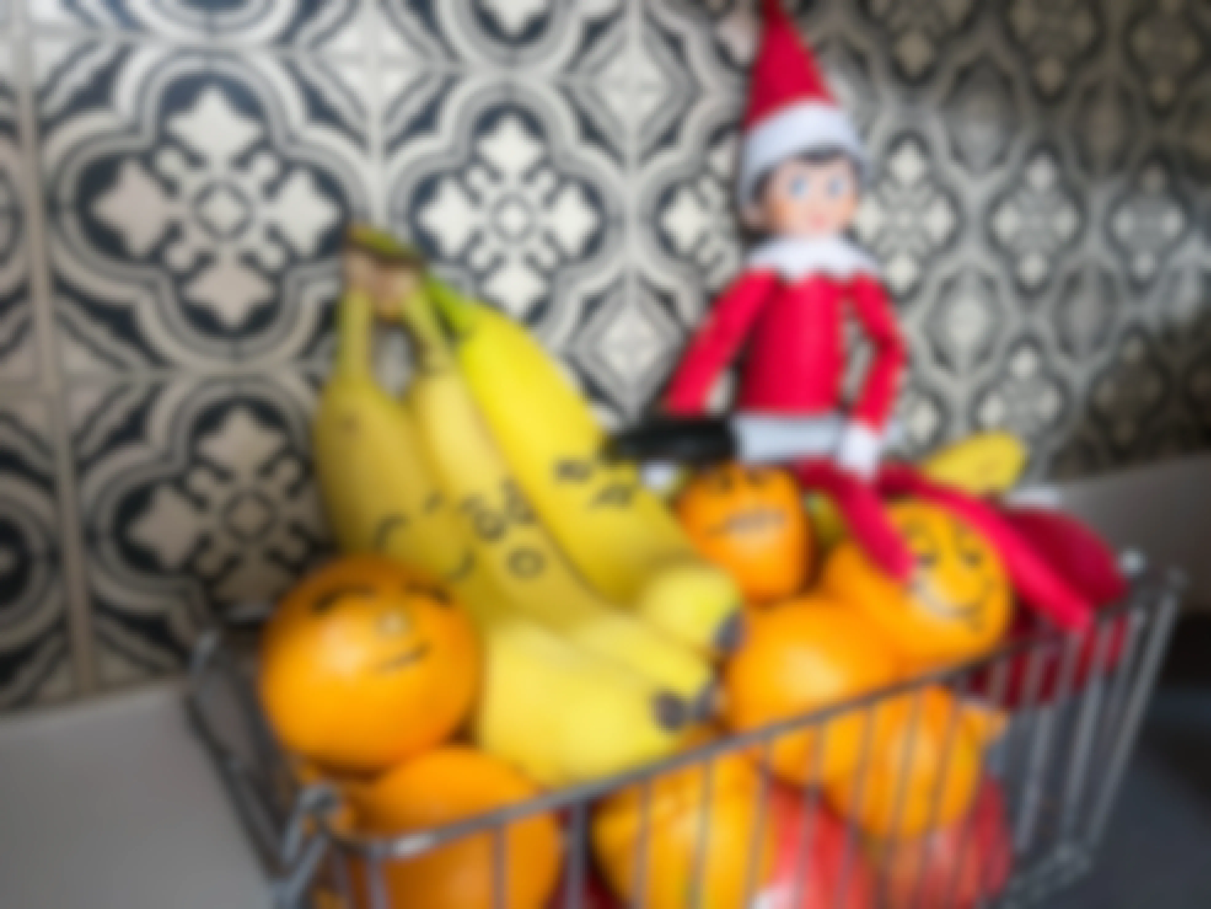 an elf on the shelf doll holding sharpie with faces marked on bananas and oranges in a basket of fruit 