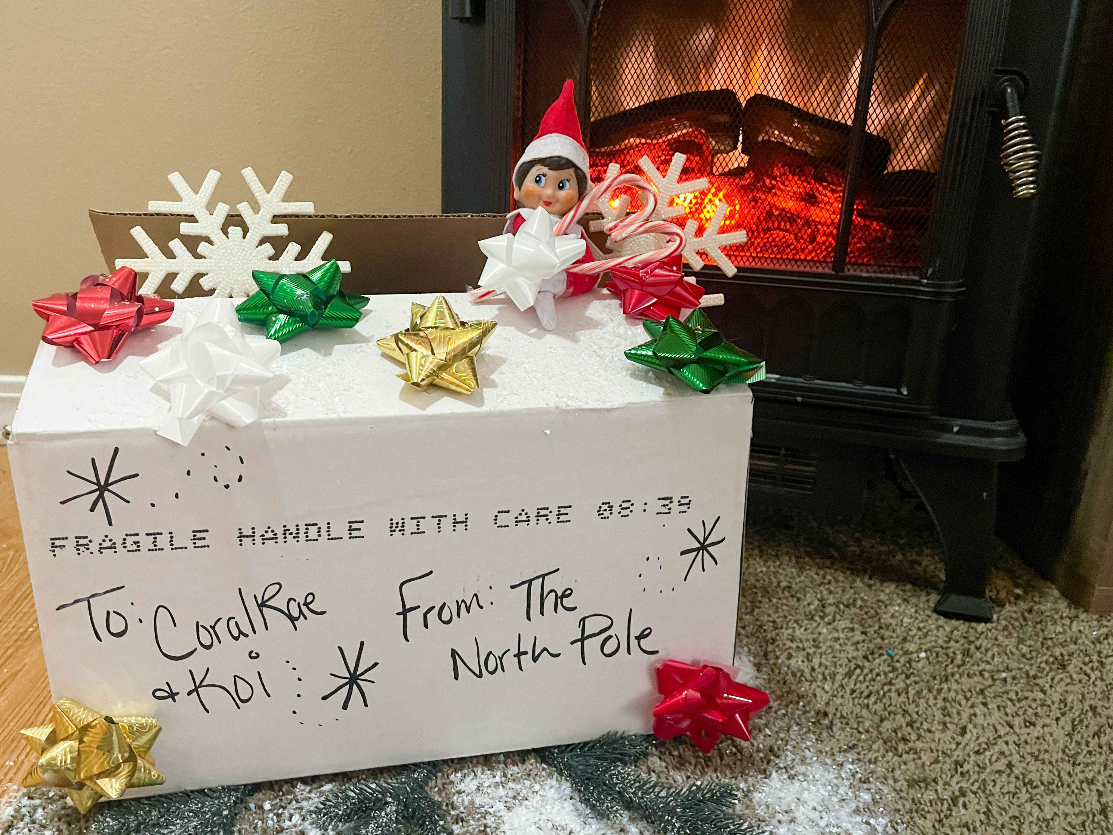 elf on the shelf doll peaking out of a box from the north pole in front of a fake fireplace 