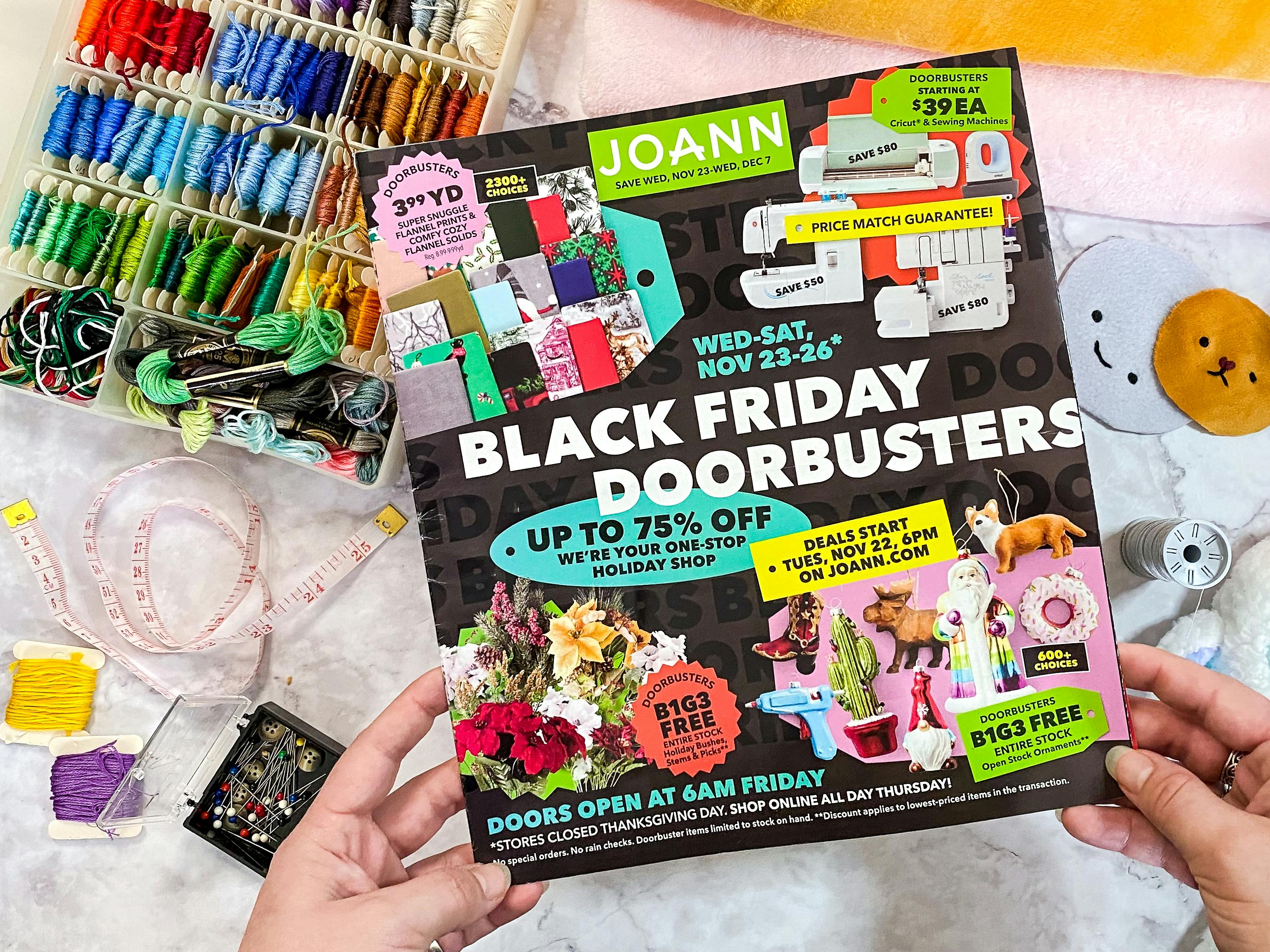Someone picking up the Joann Fabrics Black Friday 2022 advertisement from a table with sewing supplies and handmade plush dolls
