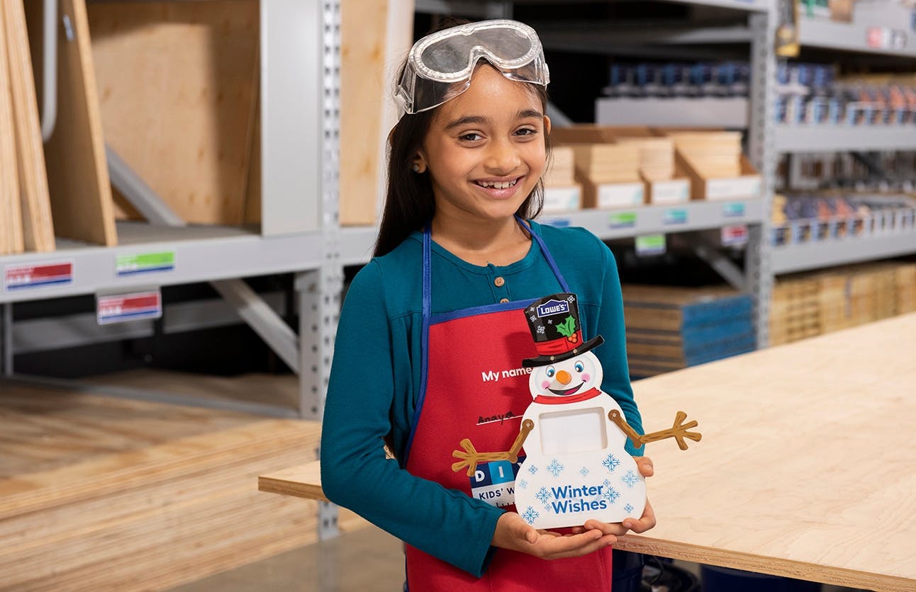 A child wearing safety goggles and a Lowe's apron holding up a snowman picture frame built using the free Lowe's kids workshop kit