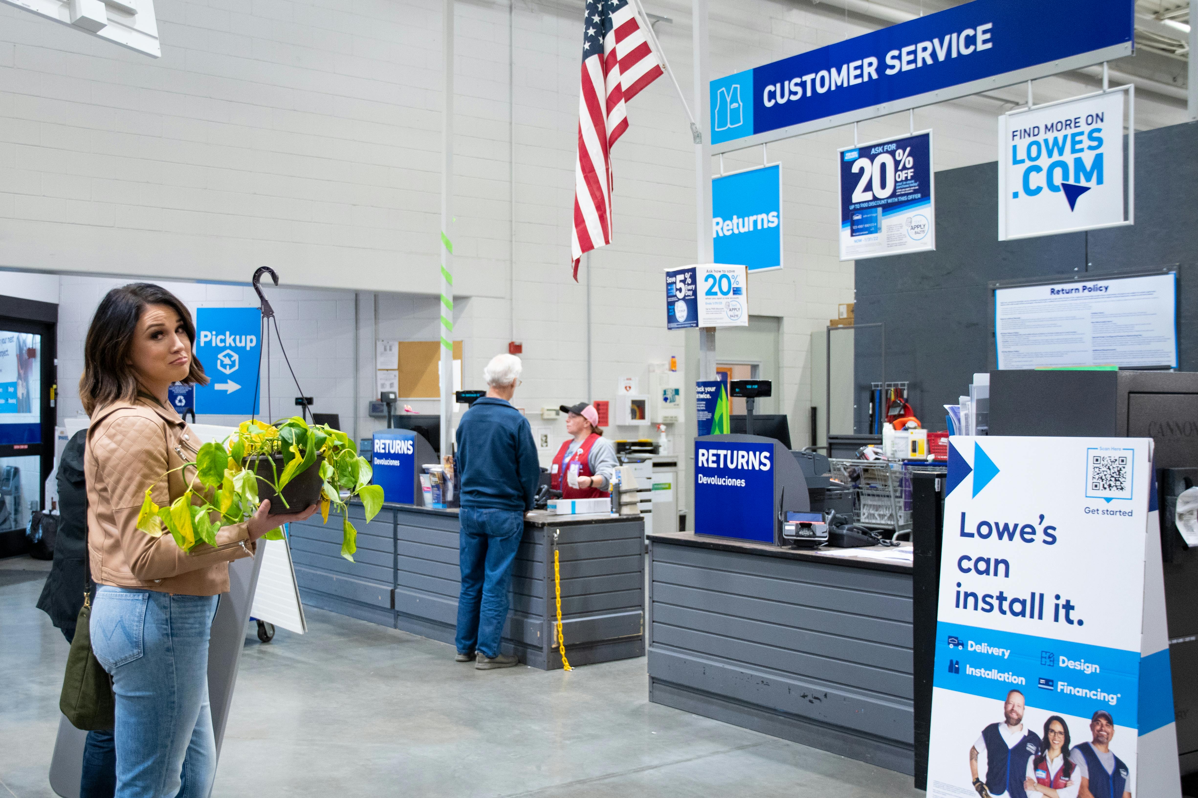 A person standing in line for the customer service desk at Lowe's, holding a dead plant.