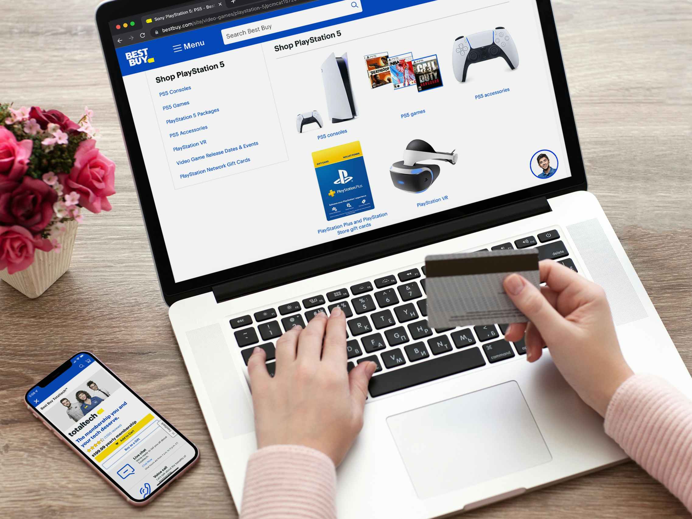 A person looking at the Playstation 5 landing page on the Best Buy website with their phone on the table next to them, displaying the TotalTech information page on the Best Buy app