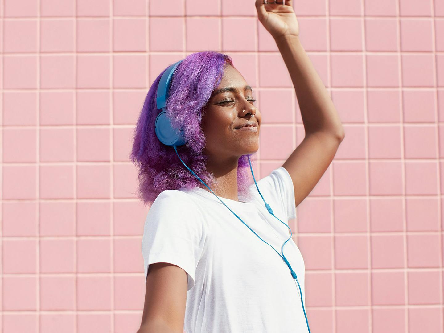 young person with purple hair, blue headphones and a white tee dances against a pink tiled background