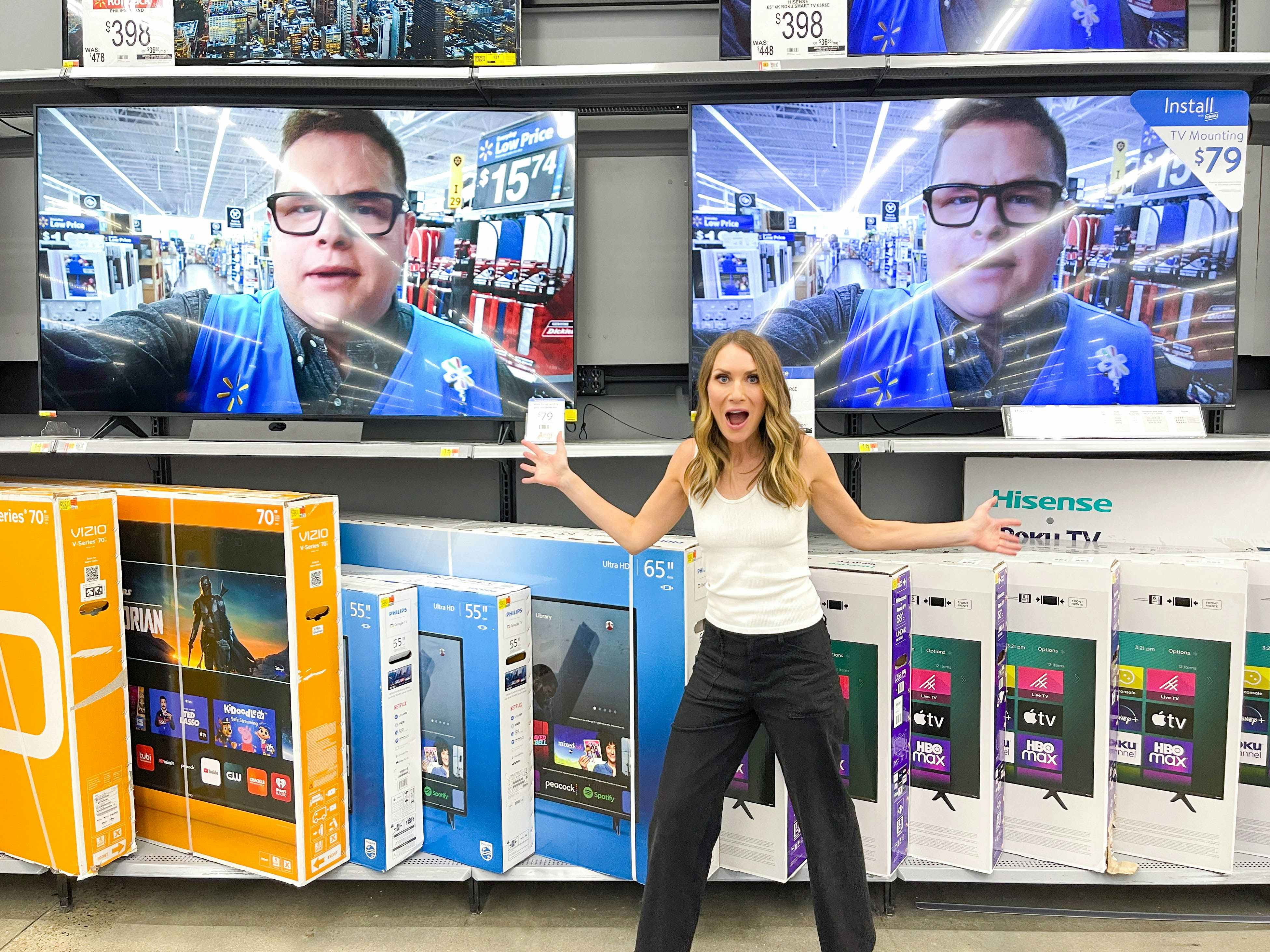 A woman standing in front of TVs on display at Walmart holding her phone and looking excited