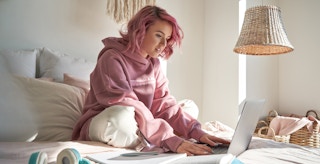 young teenager with pink hair with laptop on bed