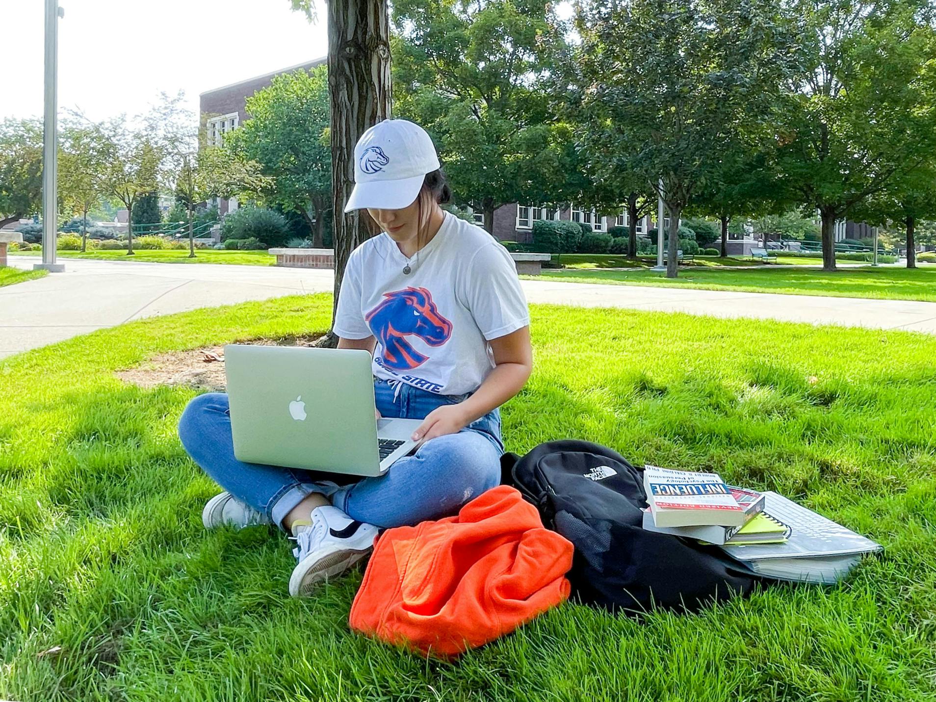 A woman sitting with books and a backpack and laptop on the lawn of a university campus.