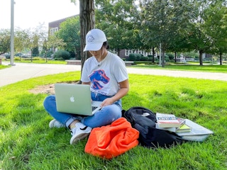 A woman sitting with books and a backpack and laptop on the lawn of a university campus.