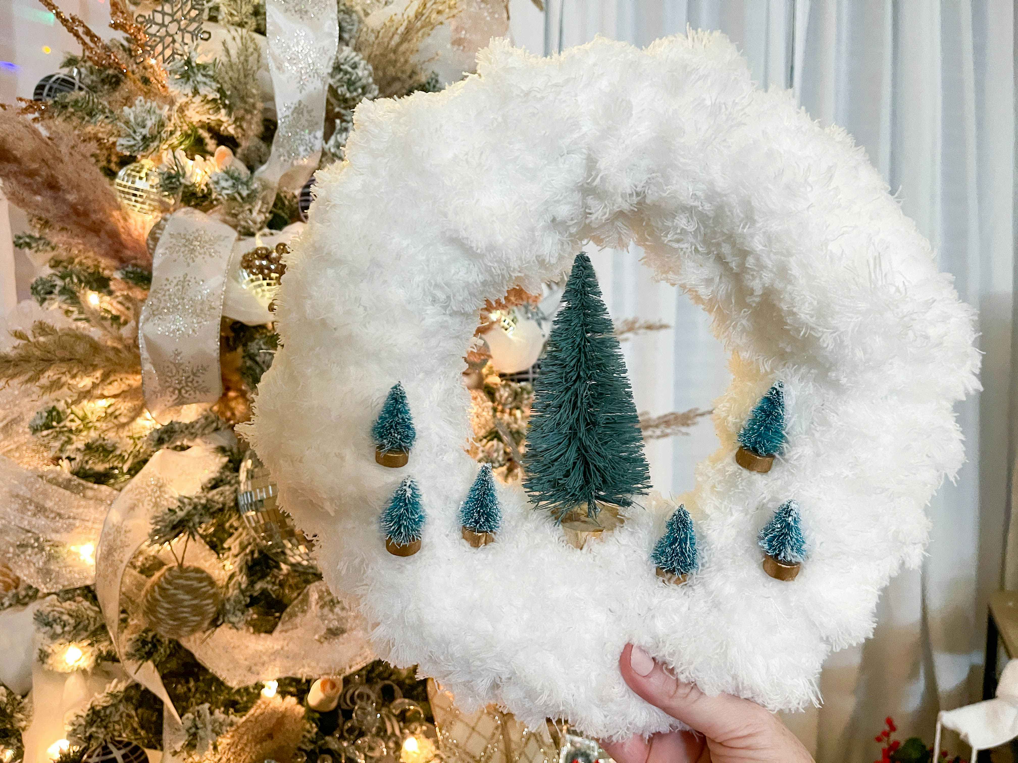 a white fluffy wreath made from mops and bottle brush trees being held in front of a christmas tree 