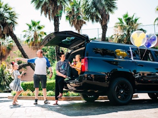 A family next to an SUV with Disney Mickey Mouse ears on and balloons coming out of a window of the car.
