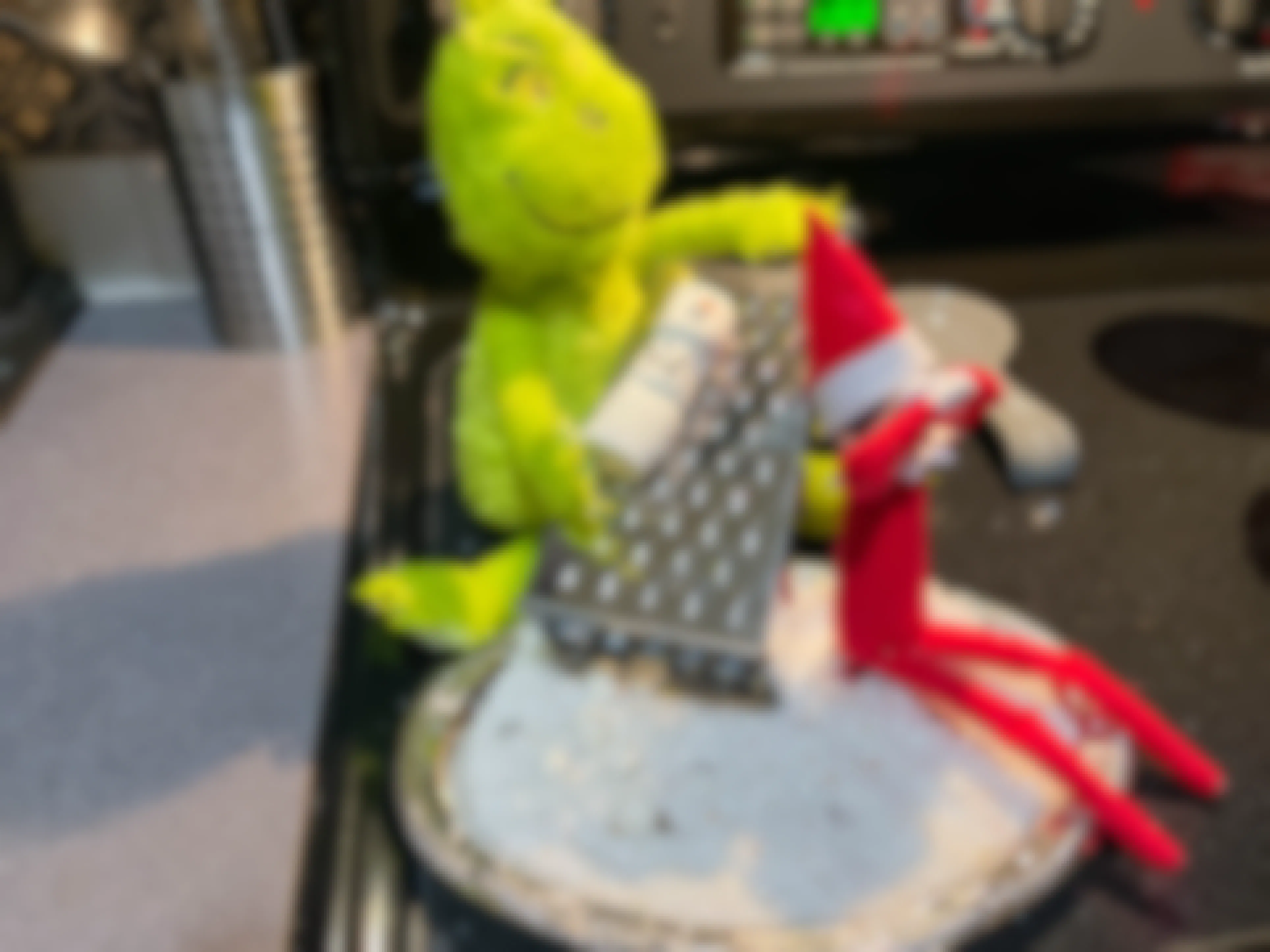 a grinch doll pretending to grate a marshmallow snowman next to a elf on the shelf doll