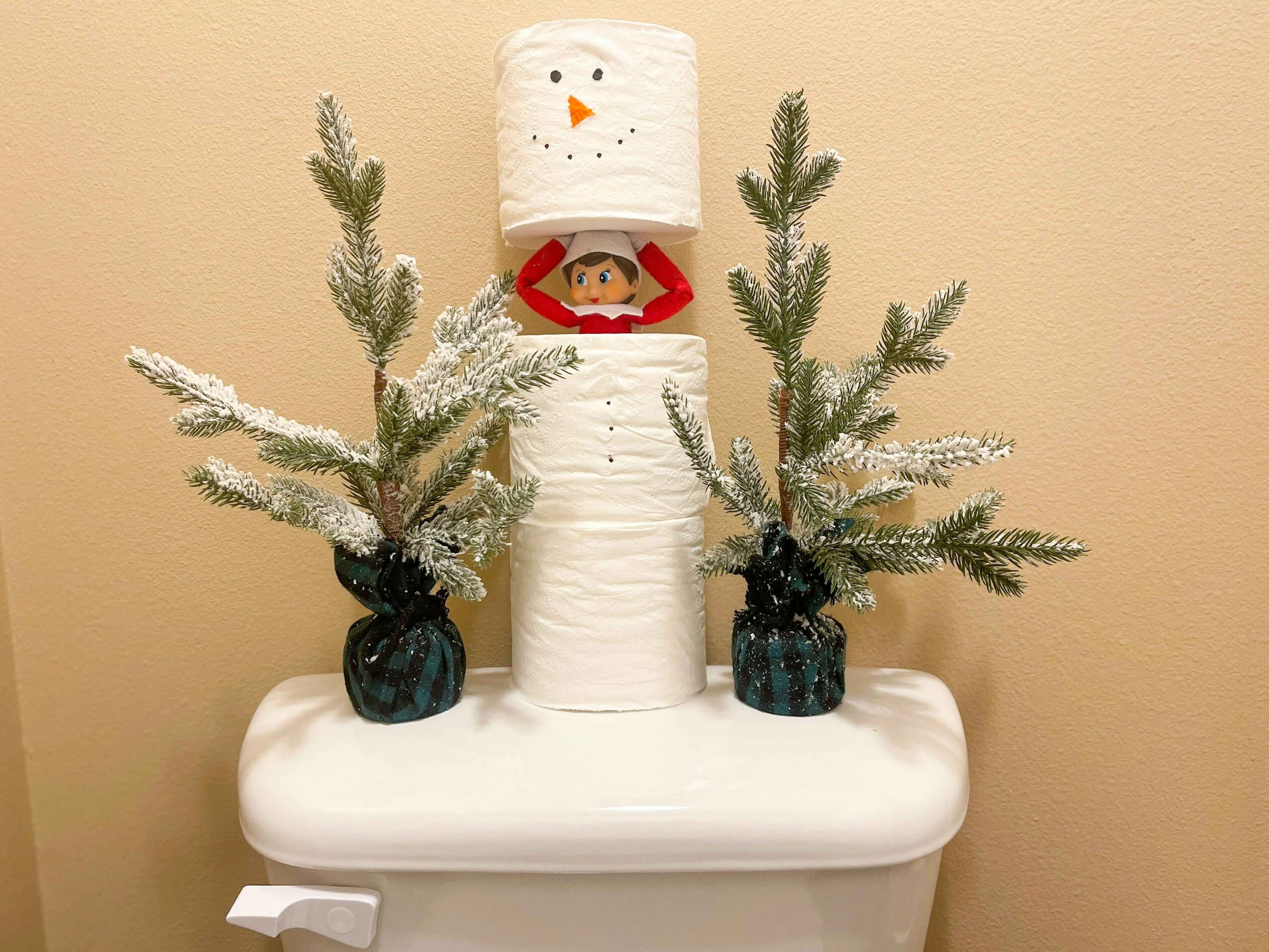 elf on the shelf hiding in stacked toilet paper made into a snowman 