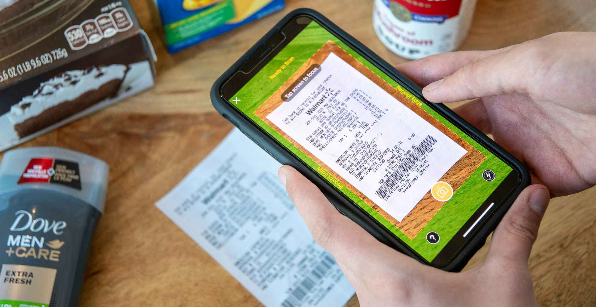A person's hands holding an iPhone with the Fetch mobile app open to the receipt-scanning feature. The user is centering the phone's camera on a receipt that is laying on a table next to some grocery items.